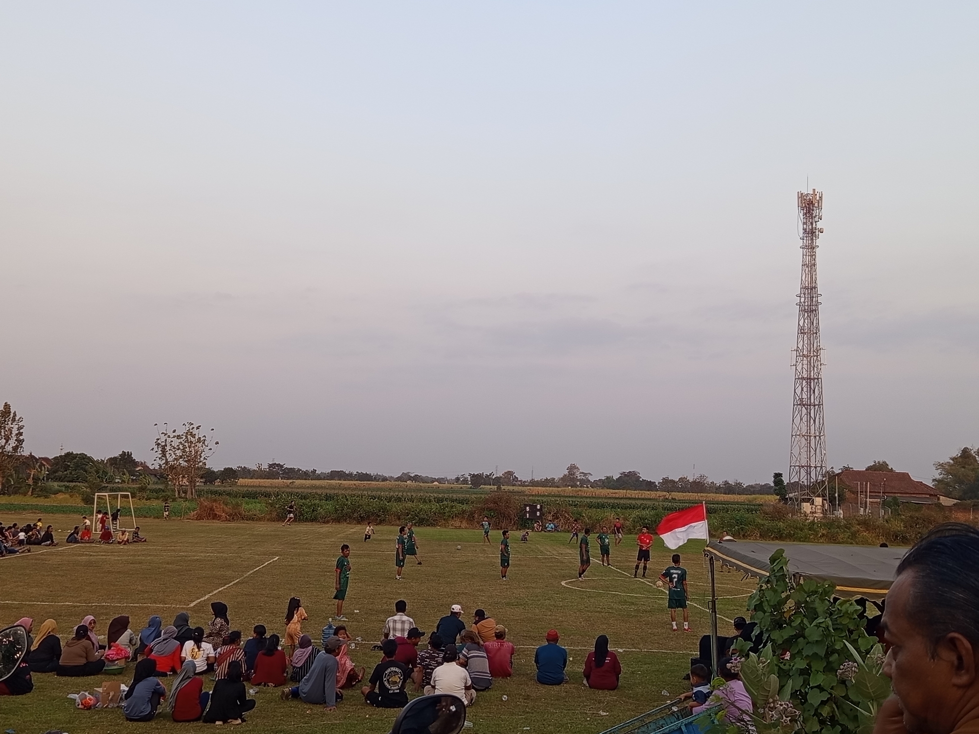 This is a medium-shot, eye-level, outdoor photograph taken at dusk in Mrican, Indonesia, depicting a soccer match being played on a grass field. The sky is a pale, hazy blue with a slight gradient. On the right side of the frame, a tall, metallic cell tower stands against the sky. A large group of spectators, primarily women and children, are seated on the grass in the foreground and middle ground, facing the soccer field. They are dressed in casual clothing, and many are wearing headscarves.
On the soccer field, several players in green and red uniforms are engaged in a game. One player in a green jersey with the number 7 on his back is prominently positioned near the right side of the field. A red and white Indonesian flag is planted on a pole on the right side of the field, fluttering in the breeze. A scoreboard is visible in the background, showing "0 1 0". The field is marked with white lines. Beyond the field, a line of trees and agricultural fields are visible, with a few buildings in the distance. The overall atmosphere appears relaxed and communal, suggesting a local sporting event.