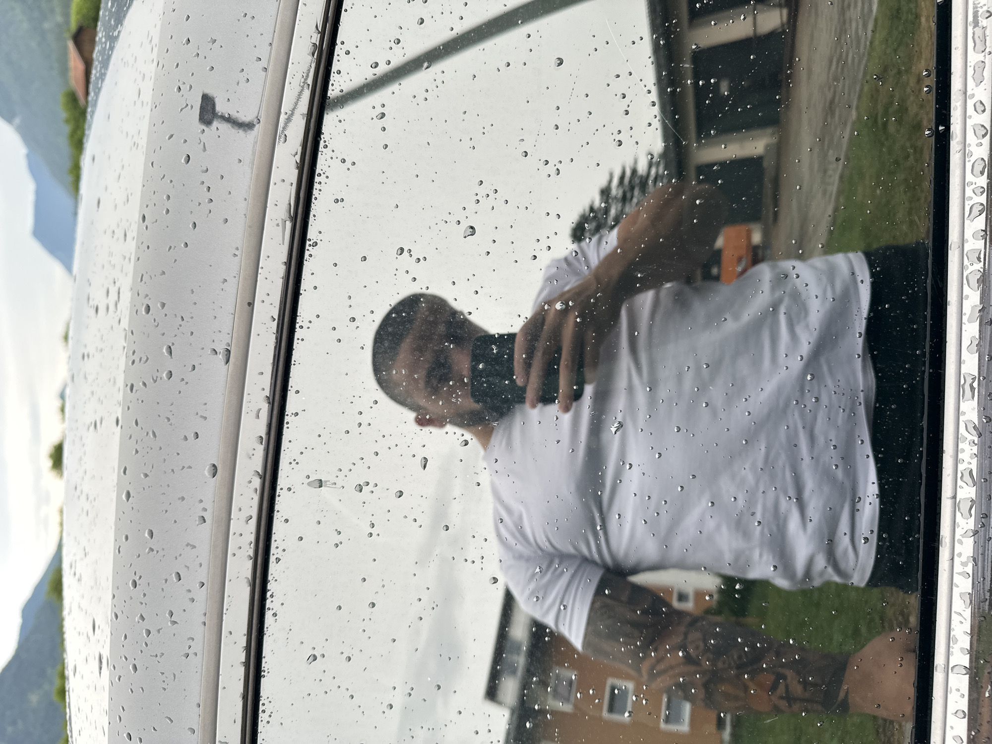 The image is a reflection on a window covered in raindrops. A man, seen from the chest up, is reflected in the glass. He has short dark hair and a beard, and is wearing a white t-shirt. His right arm, which is extended upwards, is covered in tattoos. He appears to be holding a dark rectangular object, likely a phone, in his right hand, possibly taking a selfie.
The background reflection shows a building with windows and a grassy area. The sky is overcast, suggesting cloudy weather. The presence of raindrops on the window indicates it is raining or has recently rained. The overall setting appears to be outdoors, with hints of distant mountains in the upper left corner, consistent with a location like Garmisch-Partenkirchen, Germany. The mood is neutral, captured in a moment of self-reflection. No visible text is present in the image.