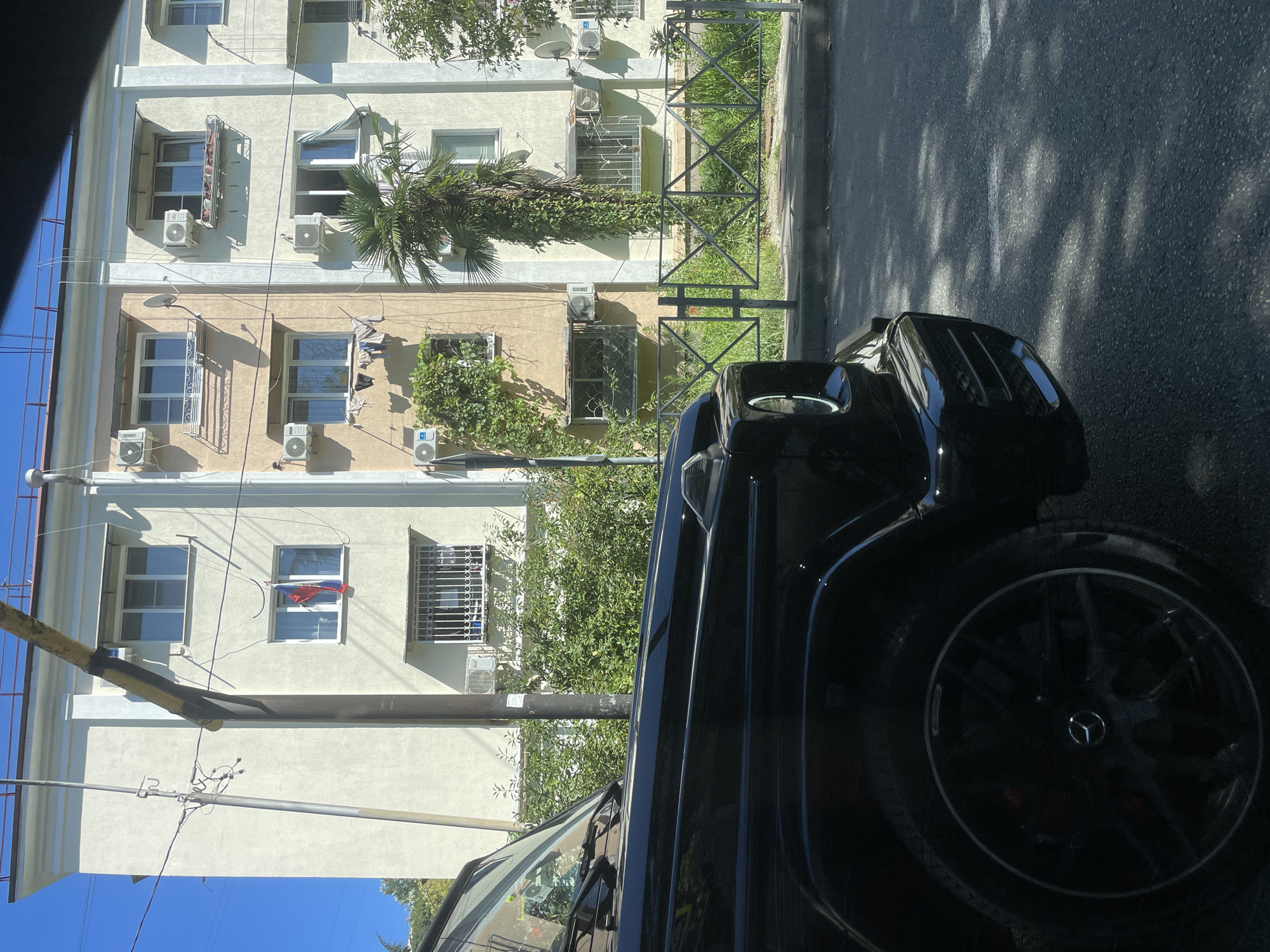 The image shows a black Mercedes-Benz G-Class parked on a street in Sochi, Russia. The car is in the foreground, partially obscuring a residential building in the background. The building appears to be multi-story with several windows, some of which have air conditioning units installed. A Russian flag is visible hanging from one of the windows. There is a fence and lush greenery between the building and the street. The sky is clear and blue.