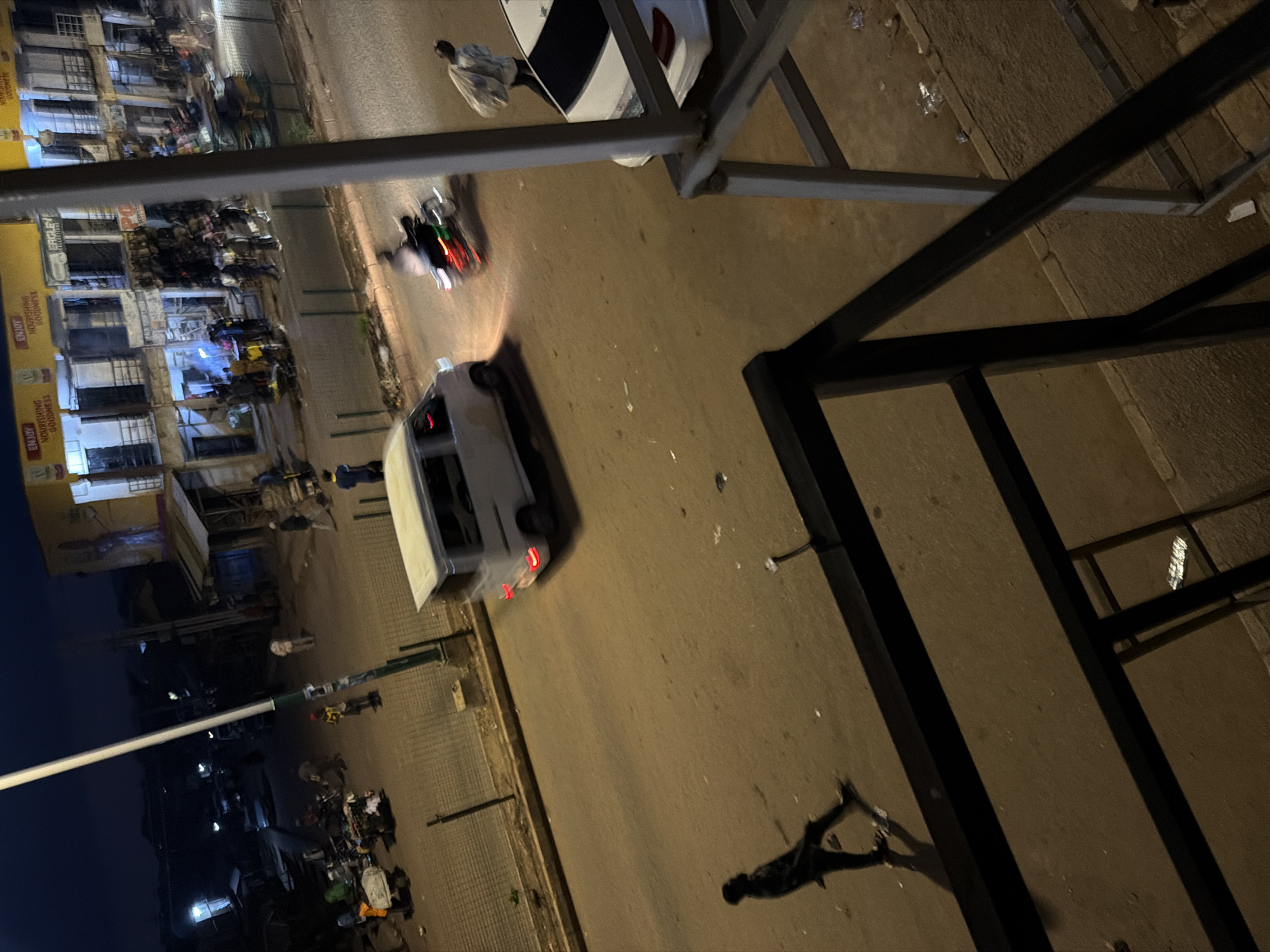 The image depicts a street scene at dusk in Bukuru, Nigeria. In the foreground, a person wearing dark clothing walks across the road, casting a long shadow. To the right, a white car is parked, and a person carrying a large bag over their shoulder walks away from the camera. Further down the street, a white van is in motion, its taillights illuminated. A motorcycle with a rider is also visible, appearing blurred due to motion. On the left side of the street, several market stalls and shops are illuminated. People are gathered around the stalls, and some are pushing carts. A tall lamp post stands prominently in the middle ground. The environment is urban, with paved roads and sidewalks, and buildings lining the street. The lighting suggests it is either late evening or early morning.