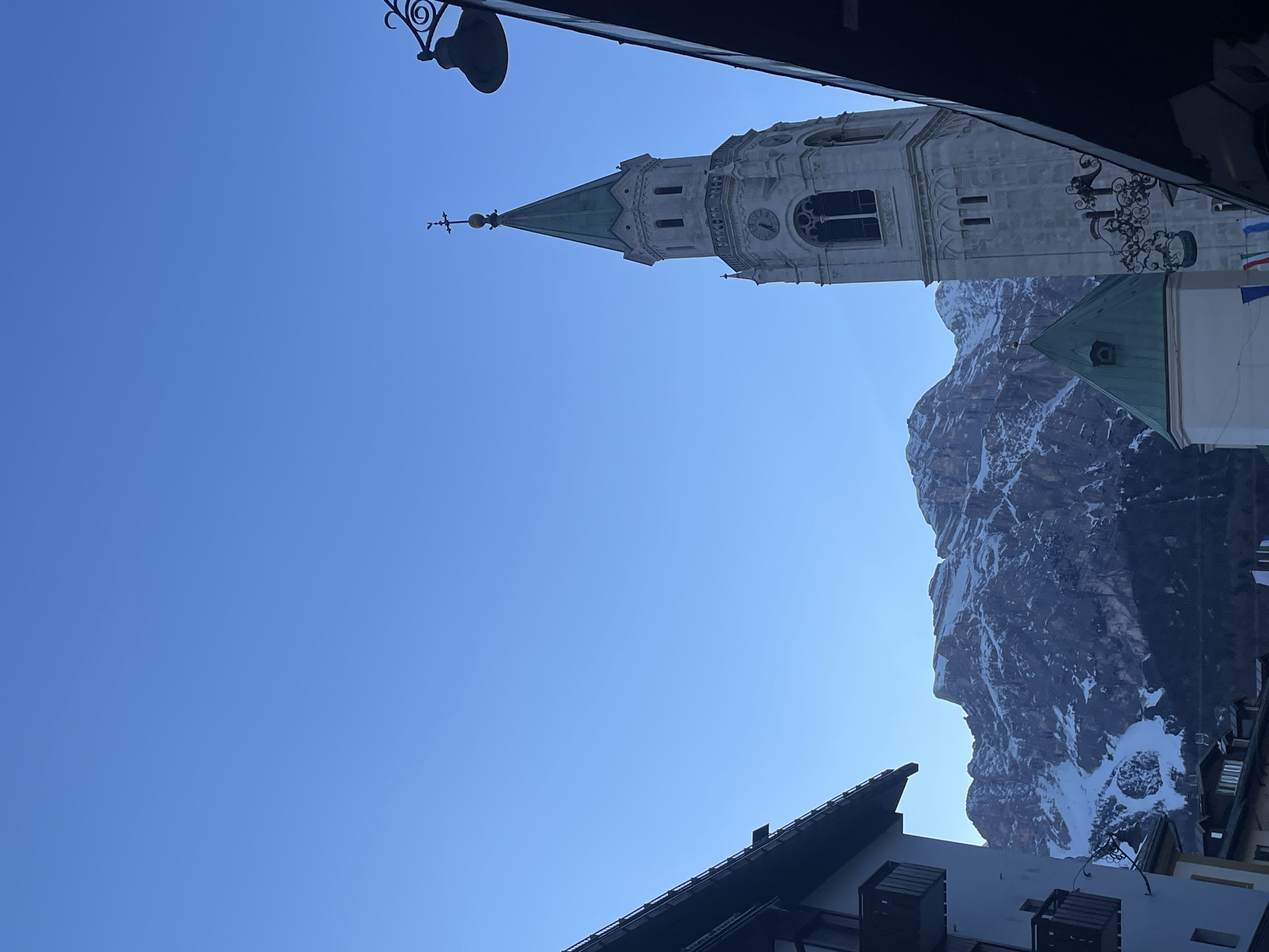 The image captures a low-angle view in Cortina d'Ampezzo, Italy, prominently featuring the bell tower of the Basilica of Saints Philip and James. The tower, crafted from light stone, rises against a clear blue sky and has a clock face visible on its facade. The upper portion of the tower narrows into a green-roofed spire adorned with a cross. A mountain range with snow-capped peaks forms the backdrop, positioned to the right of the tower. In the foreground, architectural elements, including a black wrought-iron streetlight and sections of roofs with dark eaves and white facades, frame the scene.