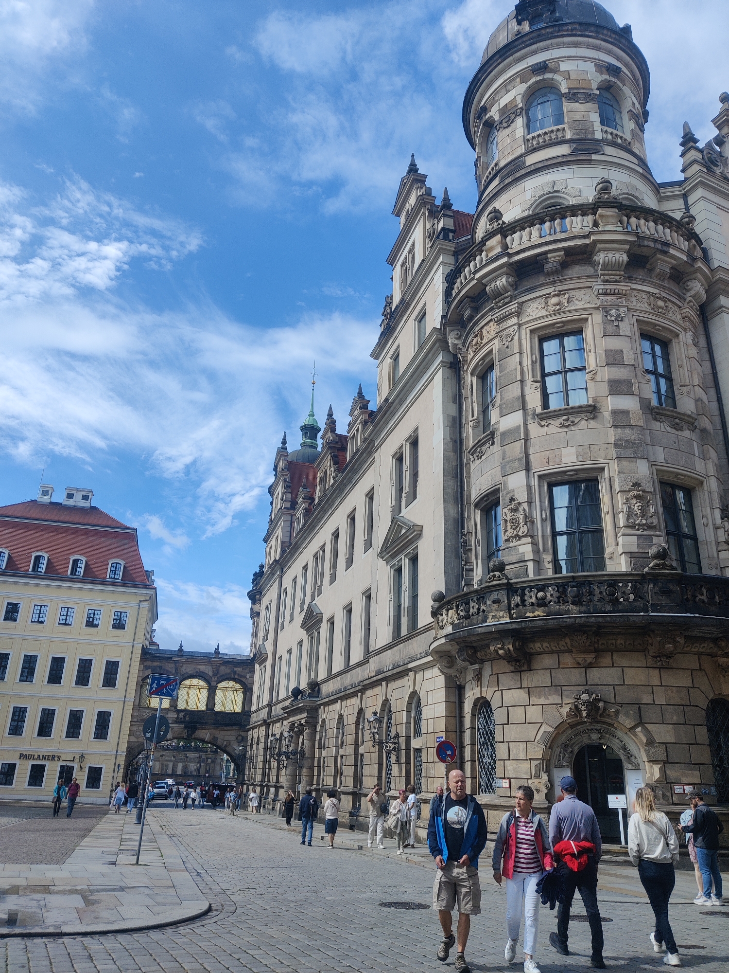 This image depicts a street scene in Dresden, Germany, focusing on the architecture of the Zwinger Palace and the Semperoper opera house. The foreground shows a cobblestone street with several people walking. A man in a blue and black jacket and khaki shorts is walking towards the camera, followed by a woman in a red and white striped shirt and white pants. Behind them, a man in a gray jacket and dark pants is walking away, and a woman in a cream-colored sweatshirt and black pants is also walking away.
The background features a grand stone building with ornate detailing, including a rounded tower topped with a dome, characteristic of Baroque architecture. The building has multiple windows and a decorative balcony. To the left, a lighter-colored building with a red roof and numerous windows is visible. An arched structure connects two buildings in the mid-ground, with a sign indicating "PAULANER'S" on the lighter building. The sky is blue with scattered white clouds. The overall impression is a sunny day in a historic European city.