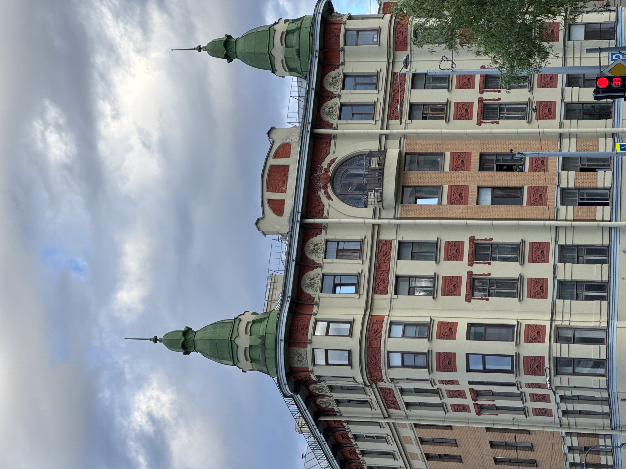 This is an eye-level, medium shot of an ornate, multi-story building in Saint Petersburg, Russia, under a cloudy sky. The building features a light-colored facade with contrasting red decorative elements and two green domed turrets topped with spires. The architecture suggests an Art Nouveau style. Windows of various sizes are visible, some reflecting the sky. The lower portion of the building transitions to a brick facade on the left. A traffic light showing red is visible in the bottom right corner, along with street signs. A tree partially obscures the right side of the building. The scene appears to be a street view on an overcast day.