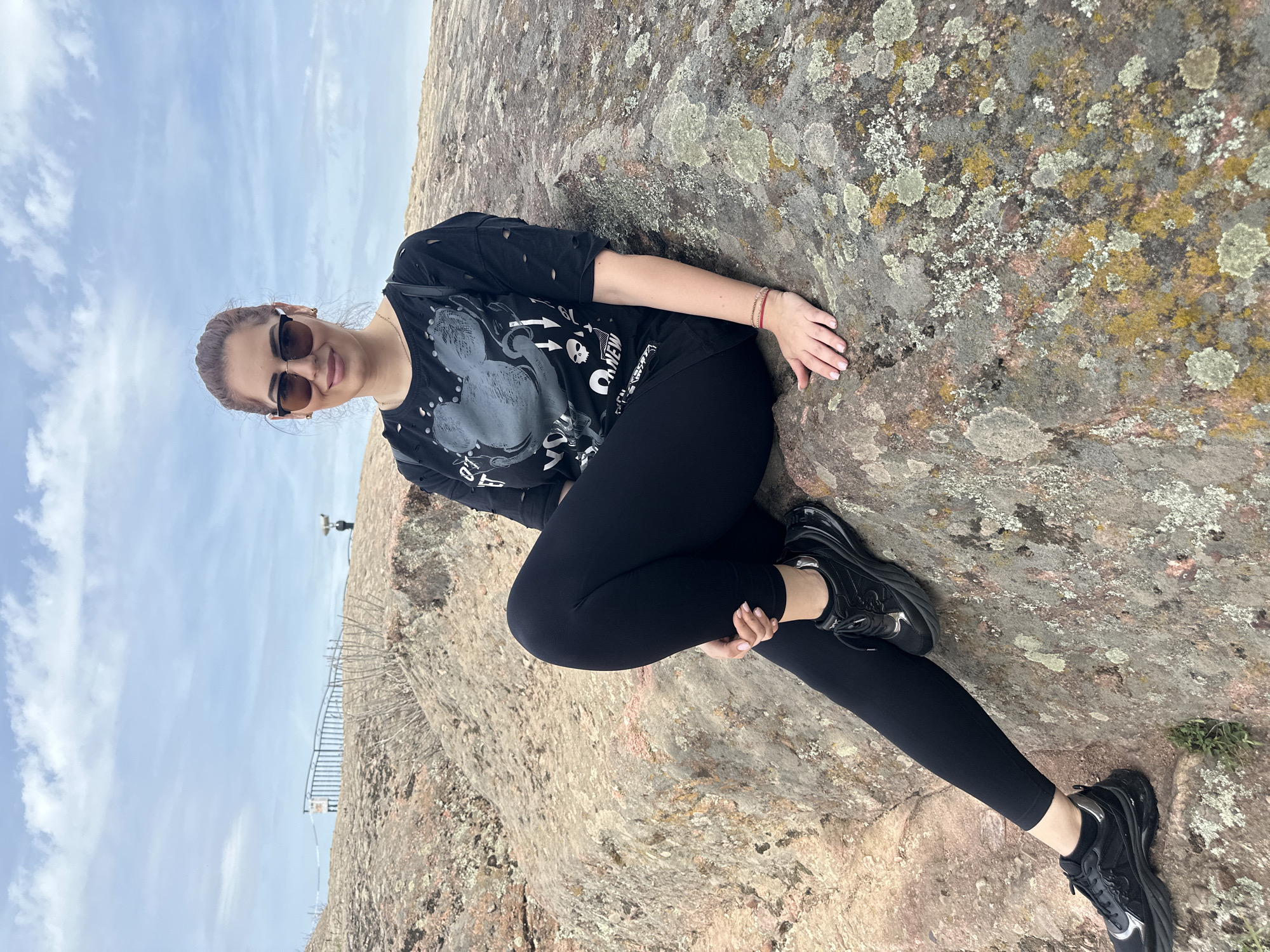 A woman sits on a rock formation in Belogradchik, Bulgaria. She wears sunglasses, a black t-shirt with a graphic print, black leggings, and black sneakers. The rock is large, with patches of lichen, and forms part of the Belogradchik Rocks formation. A railing and a distant figure are visible on another rock formation in the background, under a blue, partly cloudy sky.