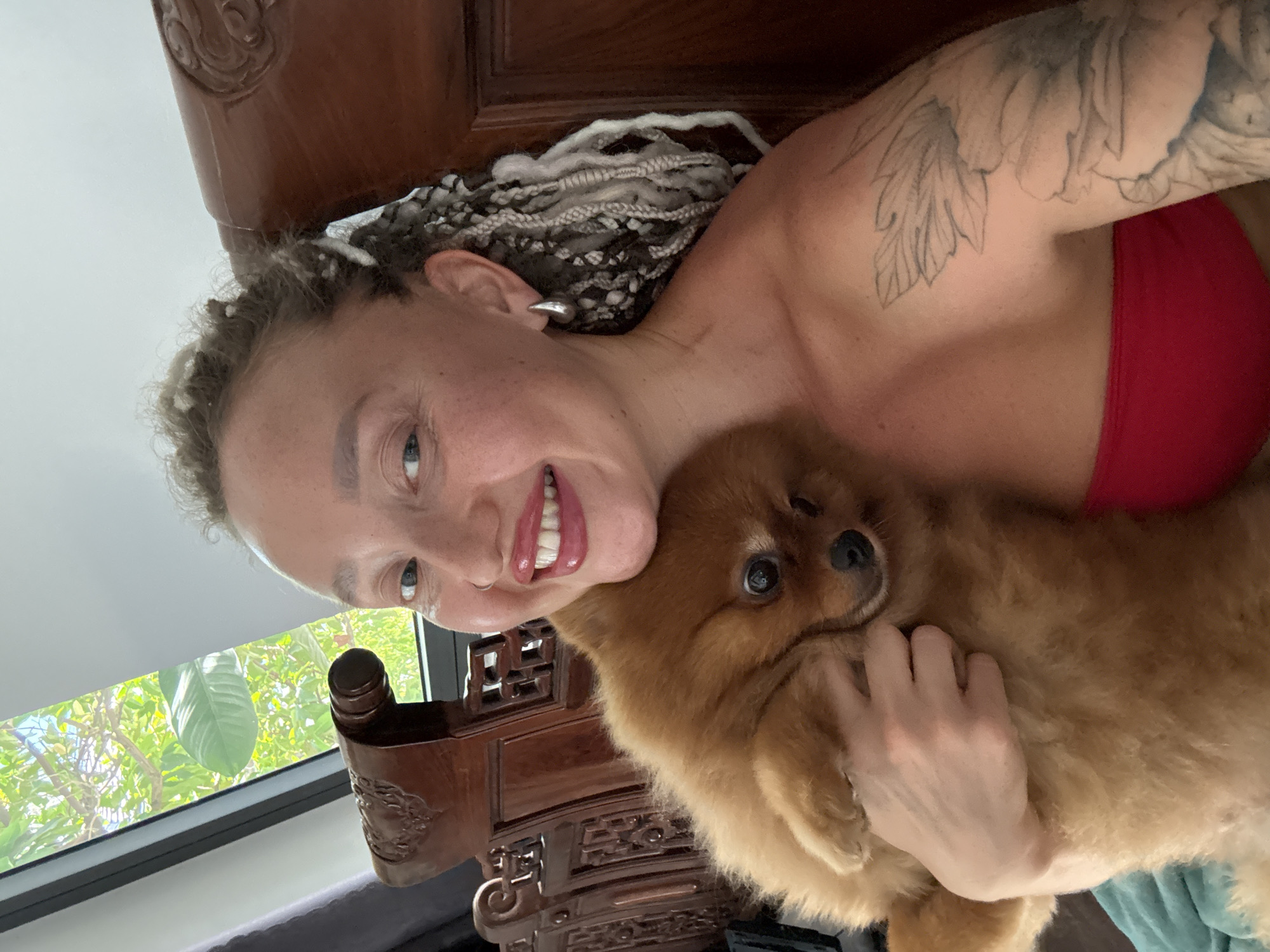 A woman with blonde hair in braids and tattoos on her arm is holding a Pomeranian dog. She is wearing a red top and silver earrings. She is smiling at the camera. The background is a wooden chair and a window with greenery outside. The image appears to have been taken indoors, possibly in a home.
