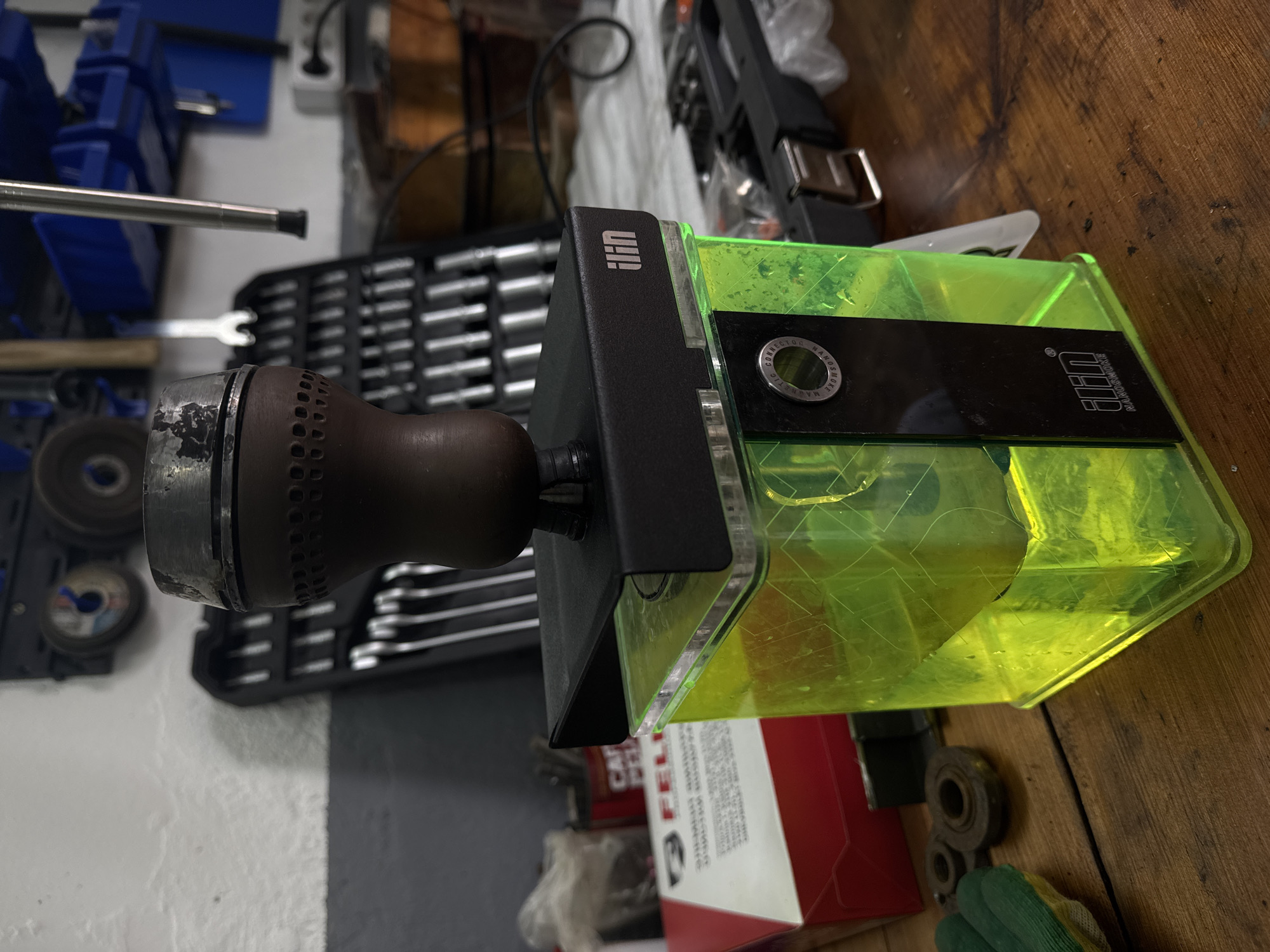 The image presents a close-up view of an object appearing to be a specialized tool, likely used in an industrial or workshop setting. The tool consists of a brown, nozzle-like component connected to a black mounting bracket. Attached to the bracket is a rectangular, neon green container or housing.
Based on the presence of tools and accessories commonly found in workshops such as wrenches, toolboxes, and abrasive disks, the location is likely an industrial workshop. There is no explicit evidence in the image to place this in the city of Chaikovsky, Russia.