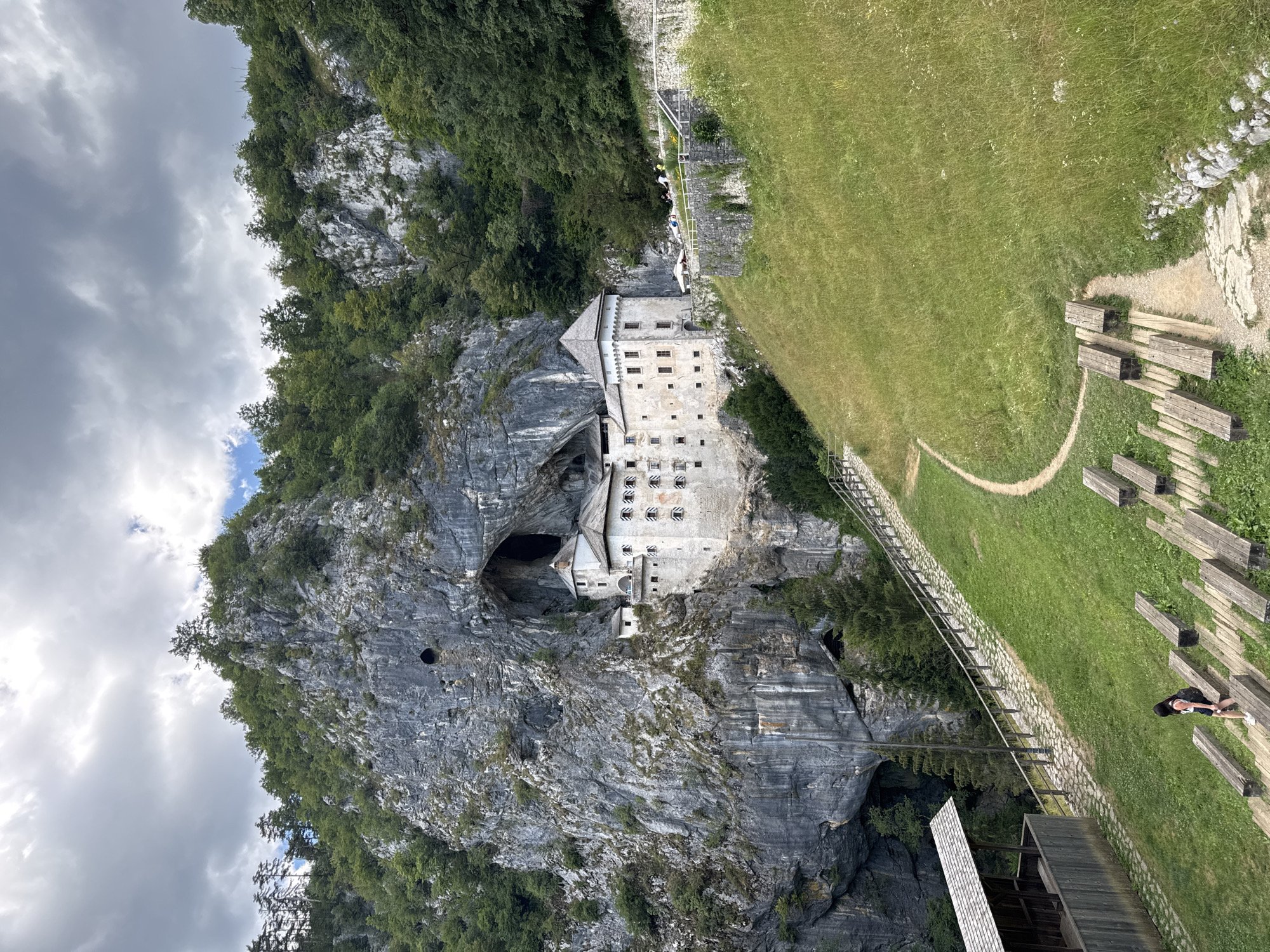 The image depicts Predjama Castle, a Renaissance fortress built within a cave in southwestern Slovenia. The castle is constructed into a sheer cliff face, with its white stone walls and multiple windows contrasting against the dark, cavernous opening behind it. The structure features several towers with conical roofs.
In the foreground, a green, grassy hillside slopes downwards. A paved pathway with a wooden railing winds up the hill towards the castle. Scattered on the grass are several wooden benches, arranged in tiered rows, suggesting an outdoor seating area or amphitheater.
A lone woman with dark hair and a backpack is walking down a stone path lined with wooden benches. She is wearing a dark top and shorts. To the left of the path, a wooden structure with a gabled roof provides shelter, possibly a waiting area or an information point.
The surrounding environment is mountainous and heavily wooded, with lush green trees covering the slopes and cliffs. The sky is overcast with dramatic, grey clouds. The overall scene conveys a sense of historical grandeur and natural beauty.
