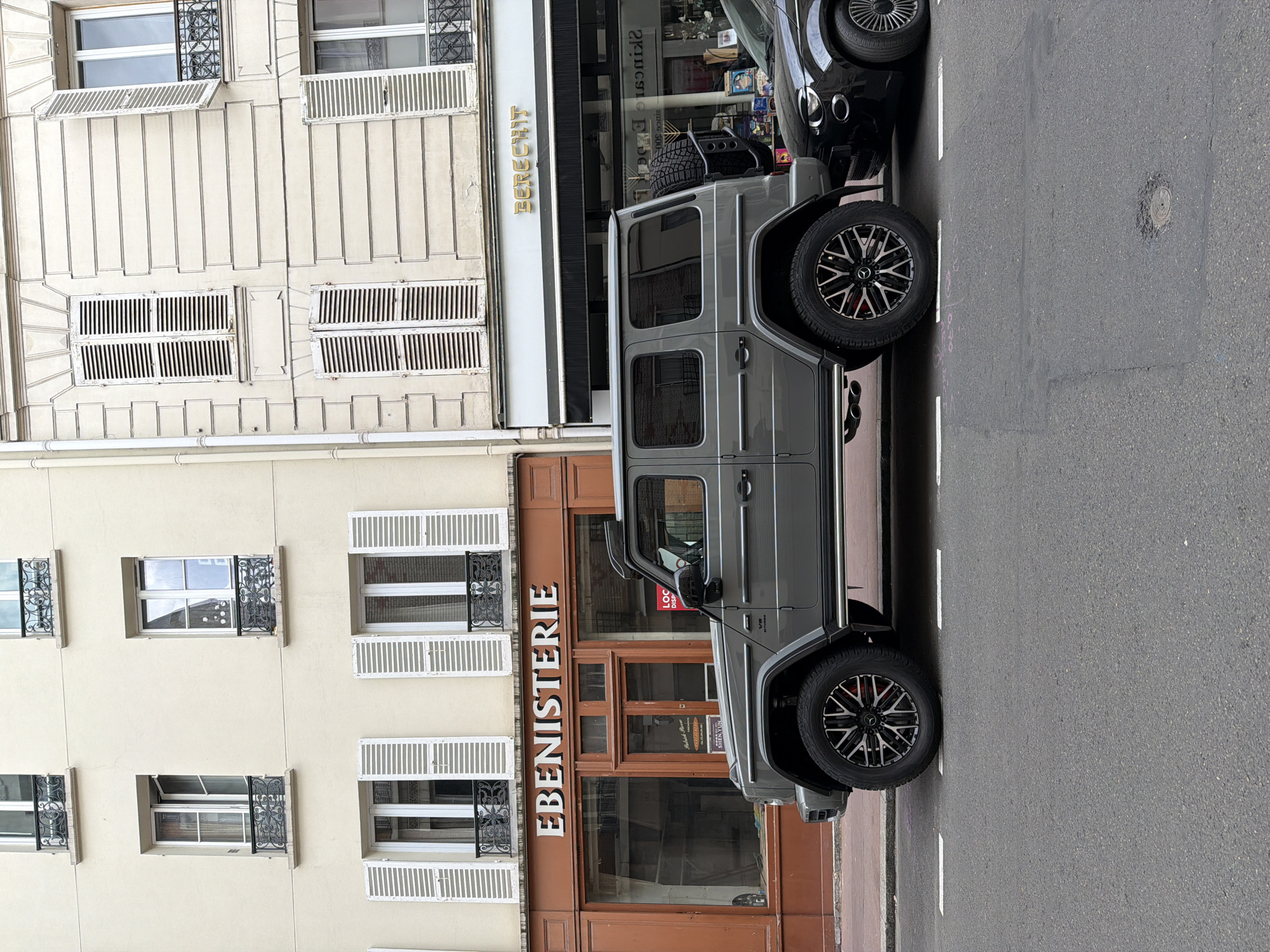 The image captures a street scene in Levallois-Perret, France. A gray Mercedes-Benz G-Class SUV is parked on the street, close to the curb. The car is positioned in front of a storefront with the word "EBENISTERIE" displayed above its entrance. The building adjacent to the EBENISTERIE has a beige facade with multiple windows, some featuring white shutters and decorative ironwork. A partial view of a black vehicle is visible behind the G-Class SUV. The street is paved with asphalt and marked with white lane dividers.