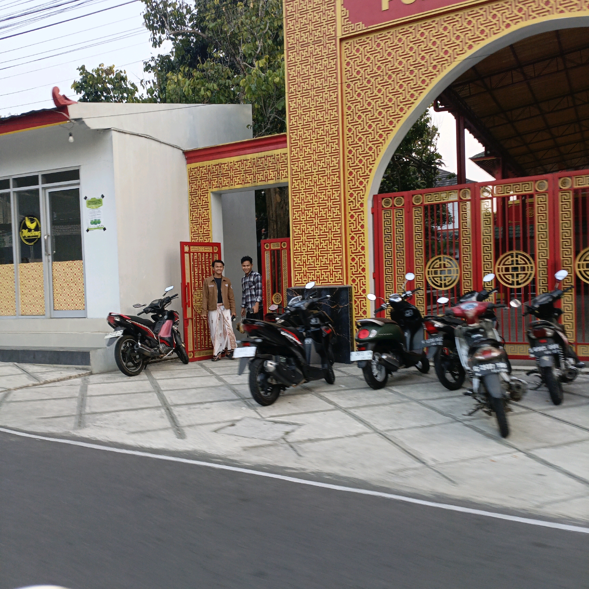 The image shows the entrance to a building with Chinese architectural elements, located on a street in Salatiga, Indonesia. A red and gold gate with intricate patterns dominates the right side of the frame. To the left of the gate is a white building with a glass door and windows, featuring a yellow circular logo with black text. Several motorcycles are parked in front of the building and gate. Two young men are standing near the entrance; one in a brown jacket and sarong, and the other in a plaid shirt. The ground is paved with concrete tiles. The overall setting suggests a commercial establishment or public building with a distinctly Asian aesthetic.