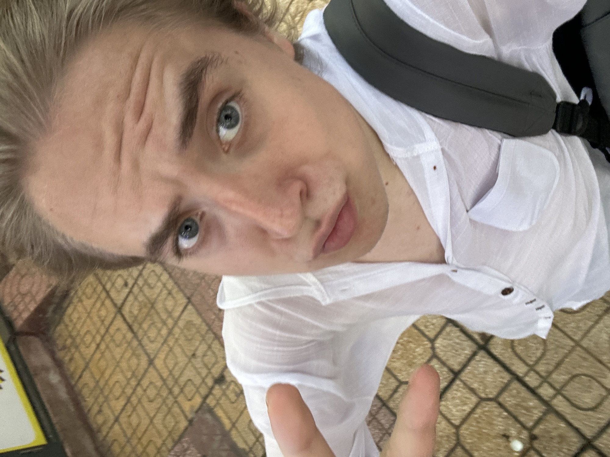 A young person, likely male, with light blonde hair and light blue eyes, is captured in a close-up, high-angle selfie. Their face is the central focus, tilted and angled upwards towards the camera. Prominent horizontal wrinkles are visible across their forehead, suggesting a raised brow or a thoughtful expression. Their lips are slightly parted.
The individual is wearing a white, somewhat sheer button-up shirt, and a dark grey backpack strap is visible over their shoulder. At the bottom right of the frame, one hand is visible, holding up two fingers in what appears to be a peace sign or "V" gesture.
The background shows a patterned ground surface, resembling sidewalk tiles or pavement with a geometric grid design, indicating an outdoor setting. The bright lighting suggests it is daytime. Based on the provided context, the scene is located in Nha Trang Ward, Vietnam. No discernible text is visible in the image.