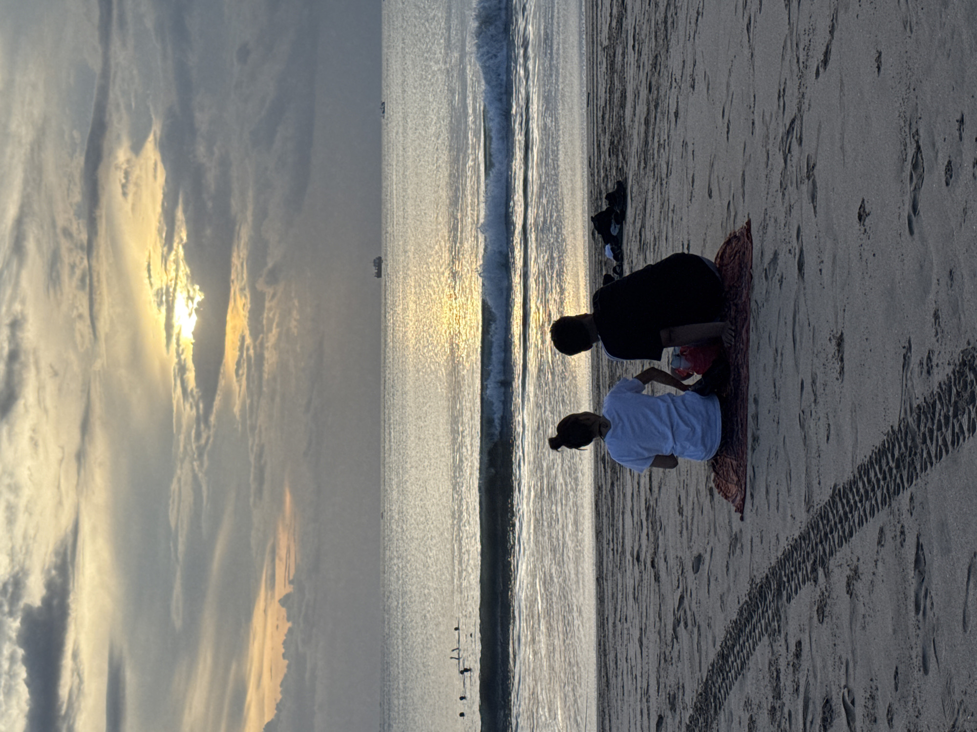 This media file captures a serene beach scene in Kuta, Indonesia, during either sunrise or sunset. The sky is partially covered with clouds, some illuminated by the low-lying sun, which casts a bright, shimmering reflection across the calm ocean surface. Gentle waves break near the shore, reflecting the sunlight.
In the foreground, two individuals are seated on a patterned mat on the sand, facing the ocean and the glowing sun. Both are seen from behind; one wears a dark top and dark bottoms, while the other wears a light-colored (possibly white) top. They appear to be observing the tranquil view, perhaps relaxing or meditating.
The sandy beach features numerous footprints and a distinct, textured tire track stretching across the mid-ground. Further back on the sand, some dark clothing items or bags are visible. On the distant horizon of the water, several small, dark shapes, possibly boats or ships, are present. Near the water's edge, a few small dark figures could be birds or debris. The overall mood is peaceful and contemplative. No visible text is present in the image.