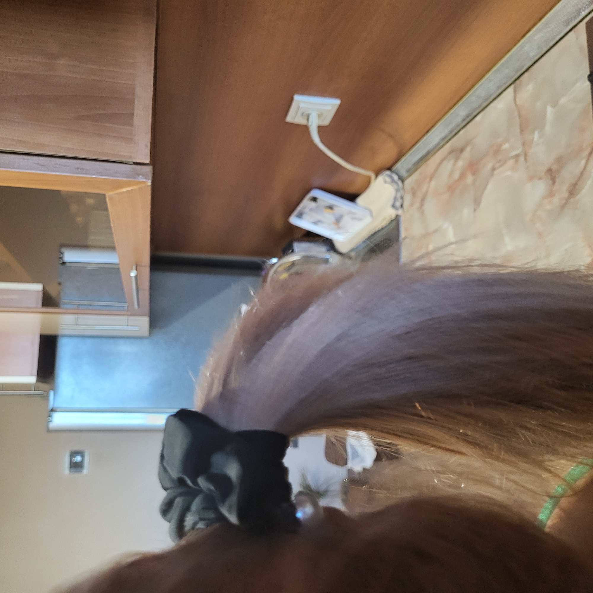 This upward-angled, candid indoor photograph primarily features the back of a person's head and hair in the immediate foreground. The person has long, light brown or auburn hair, tied back with a ruffled black scrunchie. A faint green strap, possibly from clothing, is visible on the far right.
The background shows elements of what appears to be a domestic setting, likely a kitchen or a room with cabinetry. On the left, there are wooden cabinets with a light brown finish. Above a lower cabinet section, a dark, reflective surface, possibly a stovetop or countertop, reflects light from an unseen source, resembling a window or bright light.
Further back and to the right, a warm, reddish-brown wall or cabinet panel hosts a white electrical outlet with a white cord plugged into it. Below this, a white smartphone with a light-colored case is resting, likely connected to the charger. Next to this, a section of wall features a light-colored marbled pattern, possibly a backsplash or decorative panel. In the far background, on a light-colored wall, a small, dark square object, possibly a light switch or thermostat, is visible.
The scene suggests a casual moment indoors, possibly taken from a low perspective while the person is looking upwards or moving. There is no visible text or identifiable external location cues within the image itself. The time of day is not discernible.