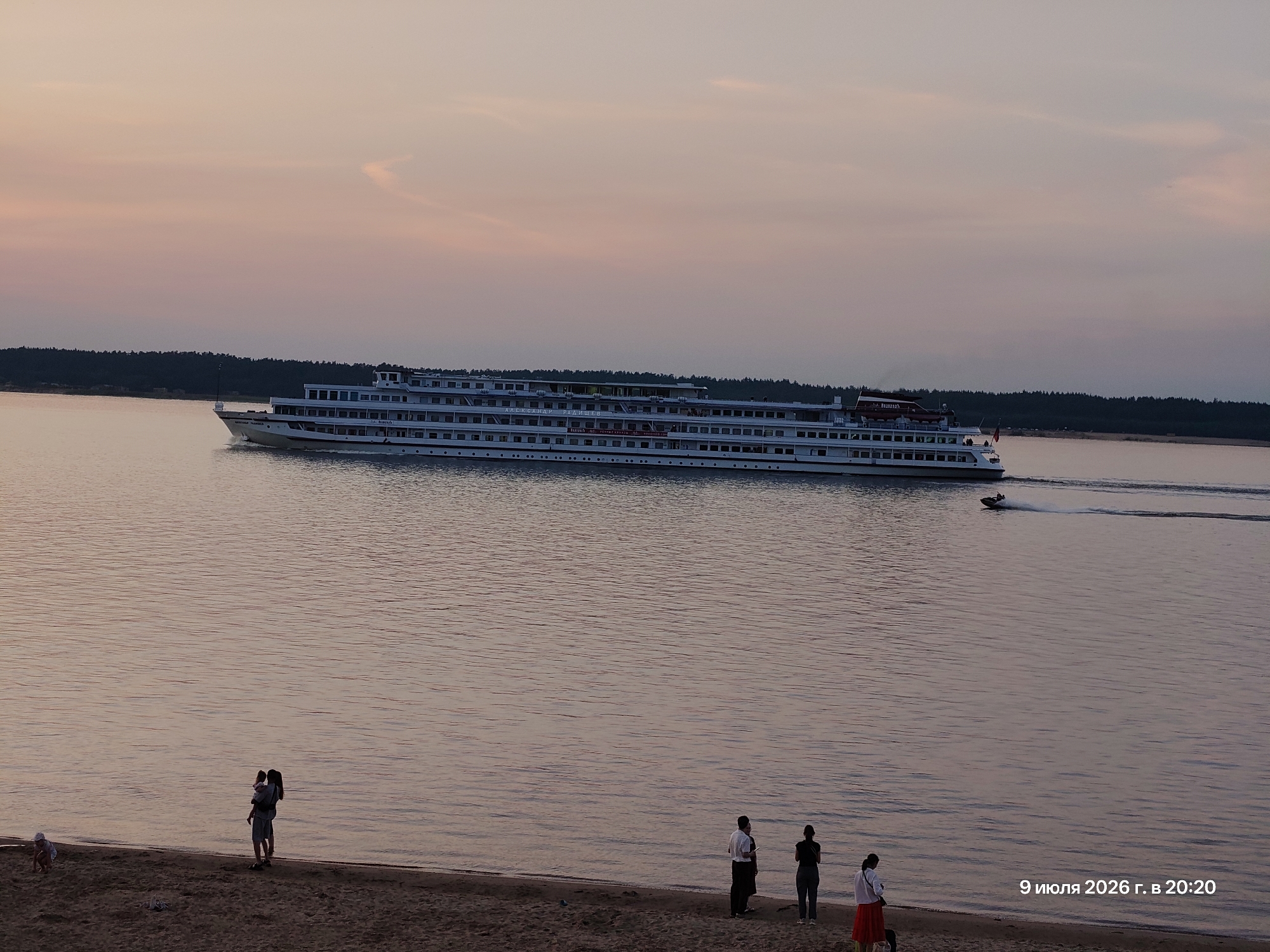 An elevated shot captures a wide river with a large white multi-deck passenger ship sailing from left to right, leaving a white wake. The ship has multiple rows of windows and appears to be moving at a moderate speed. In the distance, a dark treeline borders the far bank of the river. The sky above is a gradient of soft pink and orange, indicating either sunrise or sunset.
In the foreground, a sandy beach is visible with several people. On the left side of the beach, a child is crouched down, possibly playing in the sand. Further to the right, a group of three people stands near the water's edge. One person is holding a child, and another person stands beside them. To their right, two individuals are standing, one in a white shirt and dark pants, and the other in a dark top and pants. Further right, another person in a white top and red skirt is visible.
The overall scene conveys a peaceful, possibly evening or morning, atmosphere by the water. A watermark in the bottom right corner reads "9 июля 2026 г. в 20:20".