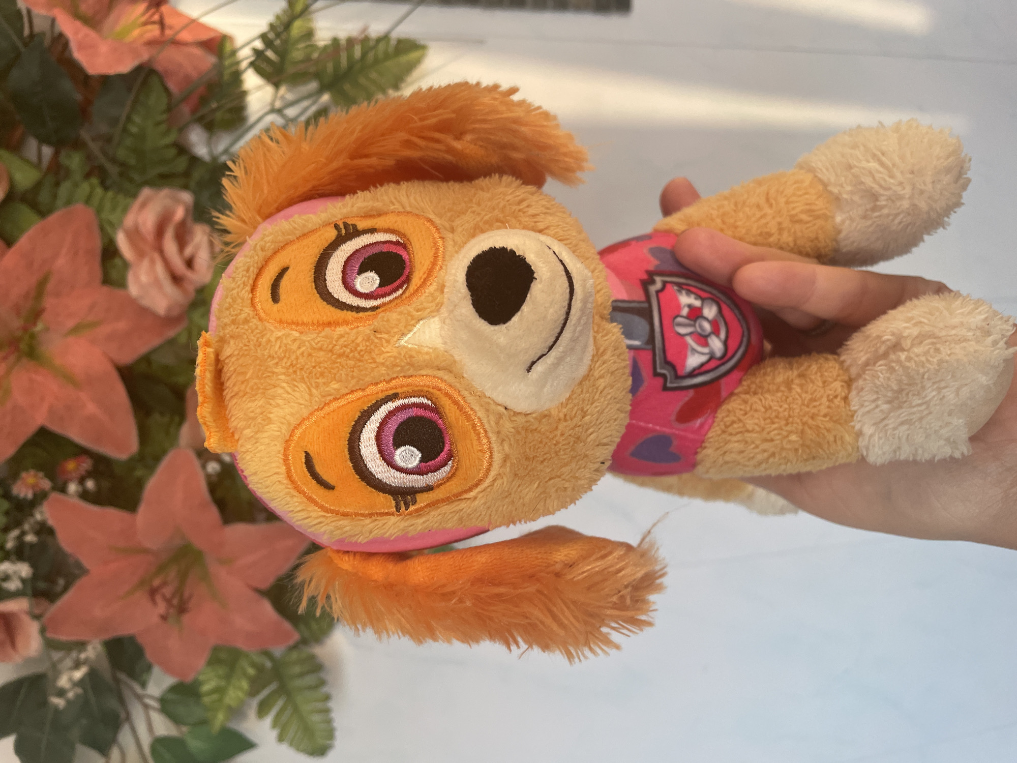 A person's hand is holding a stuffed toy dog, likely a character from Paw Patrol named Skye, in front of a white wall and a floral decoration. The dog is orange with pink accents on its ears and harness, and it has large, expressive eyes. The floral decoration behind the toy features large pink flowers and green leaves, suggesting it's part of a decorative arrangement. The lighting indicates it is daytime, with shadows cast by the toy and hand. The image appears to be taken indoors, with the white wall possibly being a background. The location is specified as Shpykolosy, Ukraine.