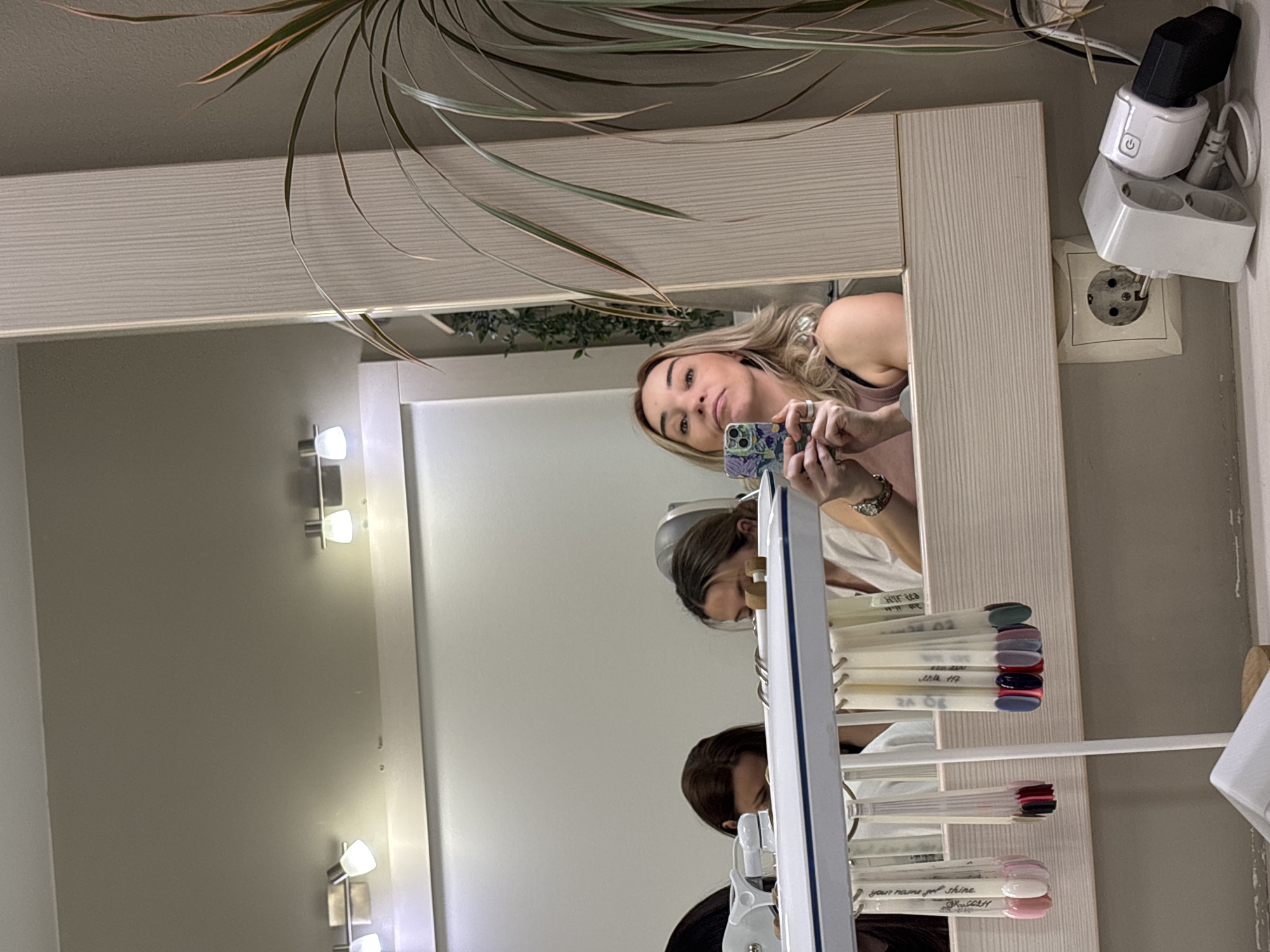 In a well-lit room, likely a beauty salon, a young woman with blonde hair is taking a selfie in a large mirror. She is wearing a dark-colored top and jewelry on her wrist and fingers. Her reflection shows her holding a smartphone with a floral case. The mirror reflects various elements of the room, including a plant with long, thin leaves hanging from above, a set of wall lamps, and electrical outlets.
The reflection also shows other individuals, possibly clients or colleagues, in the background. One person with dark hair is partially visible, and another with light brown hair appears to be looking down at something. A rack of nail polish color swatches is prominently displayed, suggesting a nail service is available in this location.
The overall scene is bright and clean, indicative of a professional establishment. The presence of a power adapter plugged into a wall socket and a surge protector indicates the room is in use and equipped with modern amenities. The image is taken from an upward angle, with the mirror reflecting the subjects from a slightly lower vantage point. The time of day is not discernible, but the artificial lighting suggests it could be any time. The location cues, such as the electrical outlets, are consistent with an indoor setting in Sinyavino, Russia. There is some visible text on the nail polish swatches, though it is difficult to read clearly.