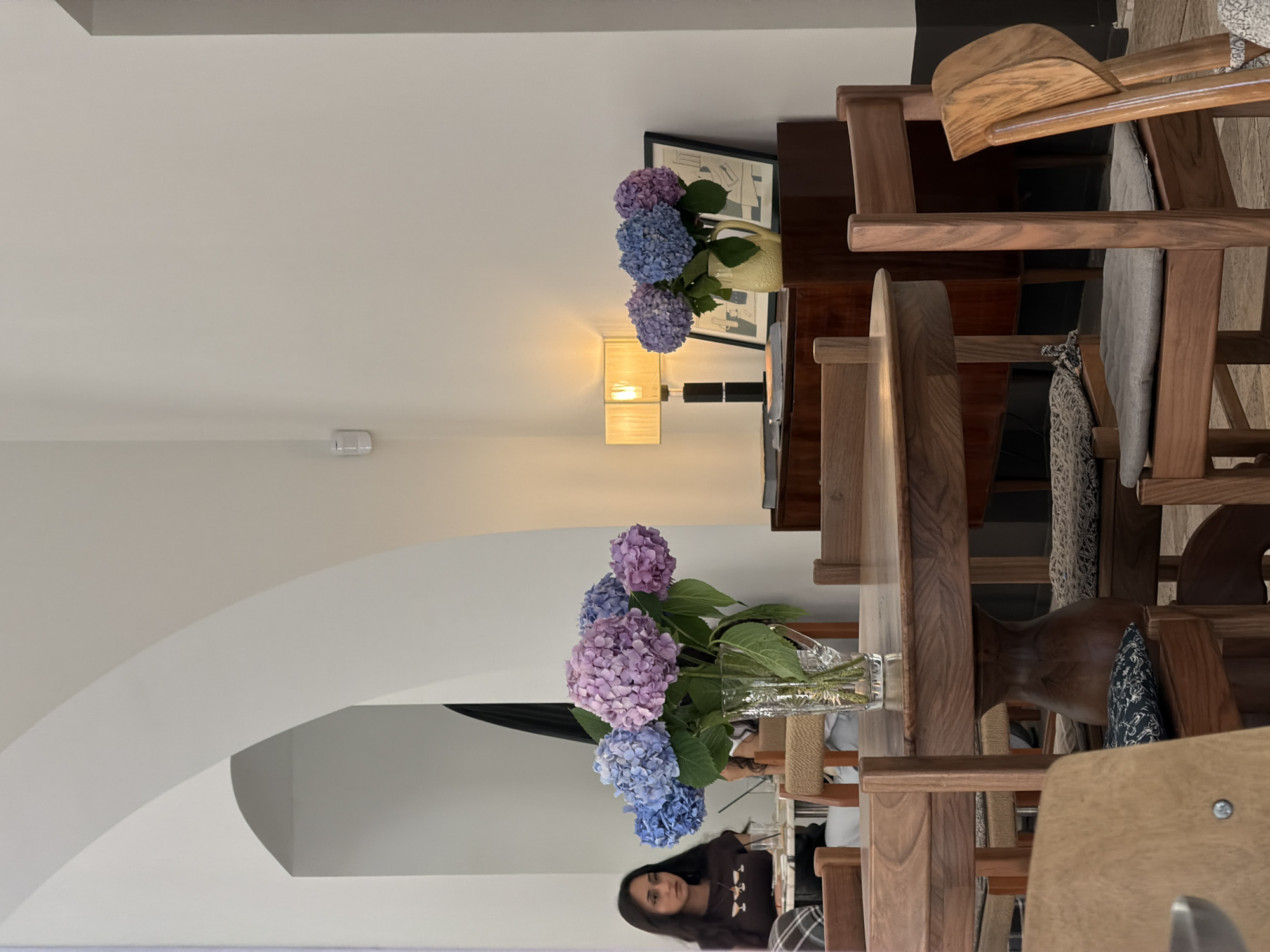The image depicts the interior of a cafe in Baku, Azerbaijan. A round wooden table is centrally placed, adorned with a clear glass vase containing a bouquet of blue and purple hydrangeas. Several wooden chairs with cushioned seats surround the table. In the background, a woman with dark hair is seated at another table, facing left, and appears to be looking down. To the right, a wooden credenza holds a lamp with a beige shade, two framed pictures, and a yellow ceramic pitcher also containing hydrangeas. The walls are painted white, and an arched architectural feature is visible on the left side of the frame. The overall ambiance suggests a peaceful and stylish cafe setting.