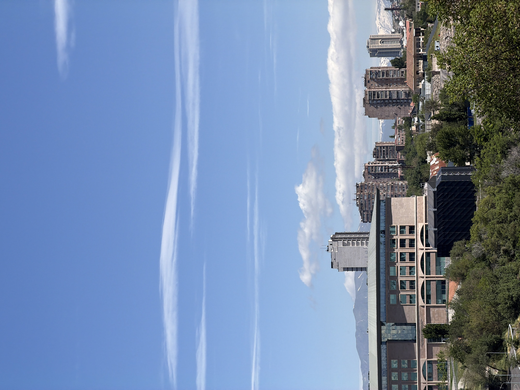 The image presents a cityscape, possibly Yerevan, Armenia, as it features architecture and terrain consistent with the region. The scene is vertically oriented, with a clear blue sky taking up a significant portion of the left side, streaked with contrails.
To the right of the sky, several buildings of varying heights and architectural styles are visible. These include older, Soviet-era apartment blocks and more modern structures. The buildings are situated on hilly terrain with some vegetation, including trees and shrubs, interspersed among them. In the distance, there are snow-capped mountains, suggesting a relatively high altitude location.
The overall impression is that of a city landscape with a mix of older and more modern architectural styles, set against a backdrop of hills and mountains. The presence of contrails in the sky indicates recent air traffic in the area.