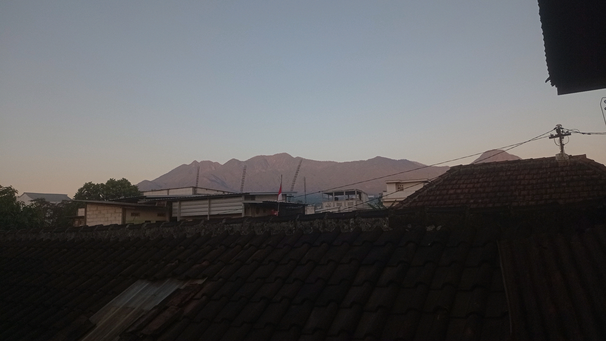 The image depicts a view of a mountainous landscape in the distance, likely seen from a residential area in Mulyoagung, Indonesia. In the foreground, several tiled rooftops are visible, some appearing aged and weathered. Beyond these rooftops, various buildings and structures are present, including what appear to be industrial or commercial buildings with corrugated metal roofs and some brick structures. Trees are scattered throughout the mid-ground, adding some greenery to the scene. In the far distance, a range of mountains dominates the horizon, their peaks partially obscured by the atmospheric haze. A utility pole with electrical wires crosses the sky. The lighting suggests it is either early morning or late afternoon, with a soft, diffused light. No people are visible in the image.