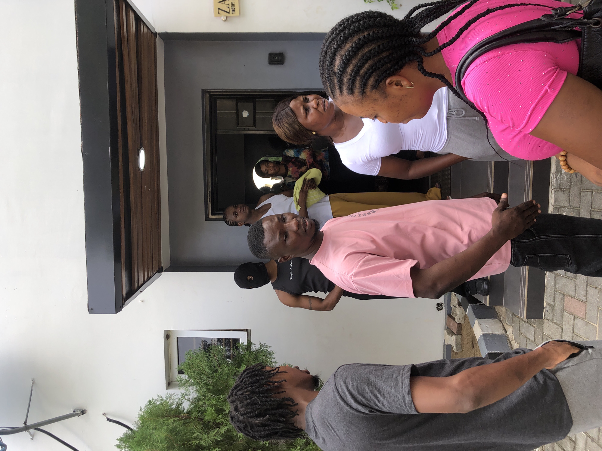 A group of seven individuals is gathered outdoors in what appears to be a daytime setting, possibly near the entrance of a building. The image is oriented vertically, showing the subjects from different angles and distances.
In the foreground, to the right, a woman with braided hair is seen from the back, wearing a bright pink, short-sleeved top adorned with small embellishments and a black bag slung over her shoulder. To her left, a woman in a white short-sleeved shirt and gray shorts stands. Both appear to be looking towards the left side of the frame. In the center, a man in a light pink t-shirt faces partially towards the camera, gesturing with his hands as if speaking. Behind him and slightly to his left, another man wears a black balaclava and a black tank top with white text, which is partially obscured. Further back, near the building entrance, are four other individuals: a man in a white tank top, a woman in a patterned short-sleeved top holding a light yellow cloth, and two other figures behind them, one of whom is also in a white top.
The setting includes a modern-looking building with a dark-framed entrance and a window. Part of the building's exterior features horizontal wooden slats and a white wall. A plaque above the doorway has visible text, reading "ZA..." and "...TIMOTHY G...", suggesting a name or address. A small, round light fixture is embedded in the wooden paneling. In the lower left foreground, a man with dreadlocks wearing a gray t-shirt is visible, looking towards the right. Green shrubbery is present near the base of the building, and the ground consists of paved interlocking bricks. The clear, bright lighting indicates it is daytime.