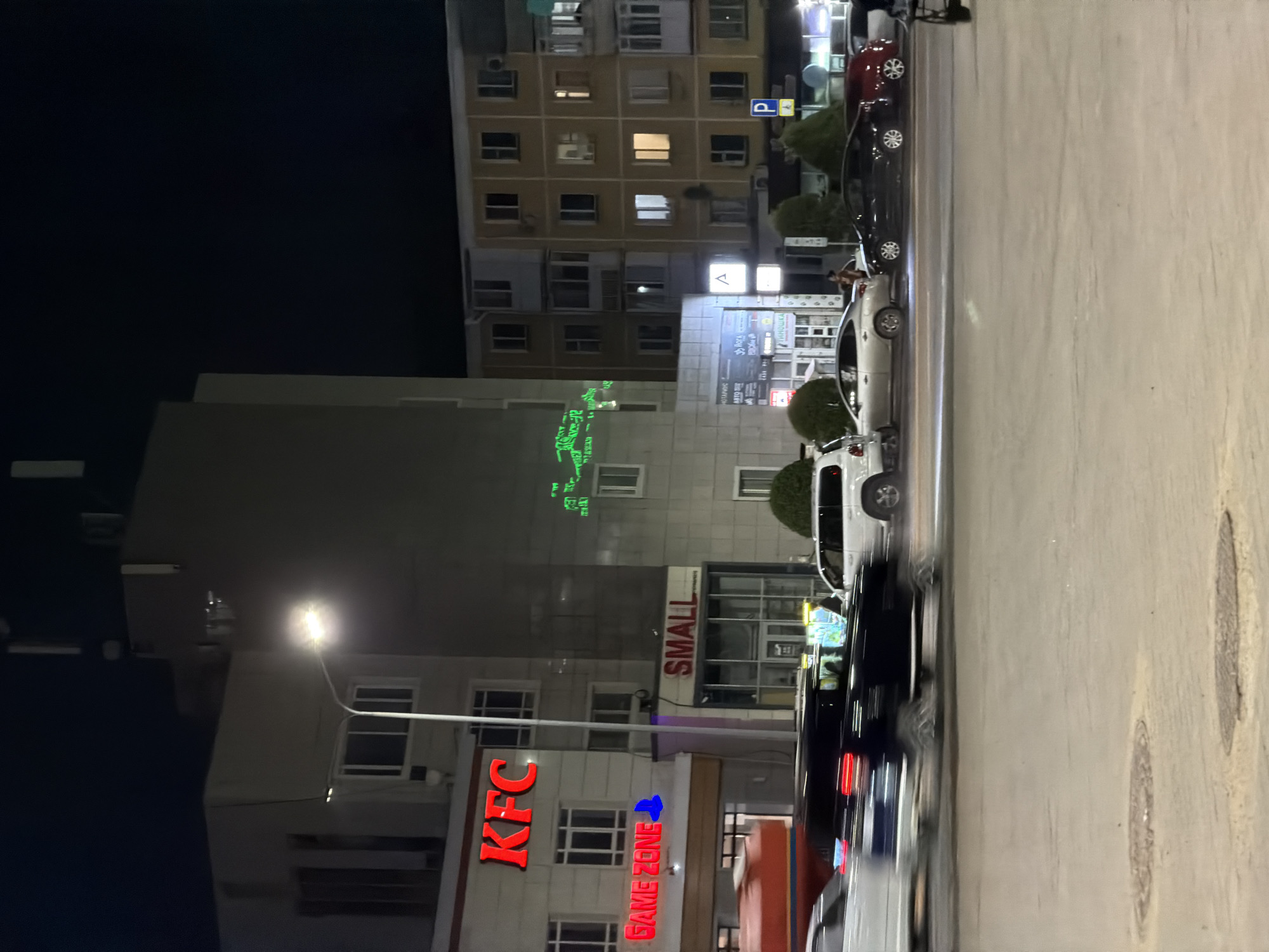This is a nighttime photograph of a street scene in Astana, Kazakhstan. The main focus is on a multi-story building with several businesses on the ground floor. A large, red KFC sign is prominently displayed on the upper facade of the building. Below it, a sign reading "GAME ZONE" with a blue icon is visible. To the right of KFC, another sign reads "SMALL".
In front of the buildings, a street is visible with several cars in motion, indicated by motion blur. The cars include a white SUV, a dark-colored sedan, and a red SUV. Streetlights illuminate the scene, casting light on the buildings and road. The sky is dark, suggesting it is late at night. The overall impression is that of a busy commercial area at night.