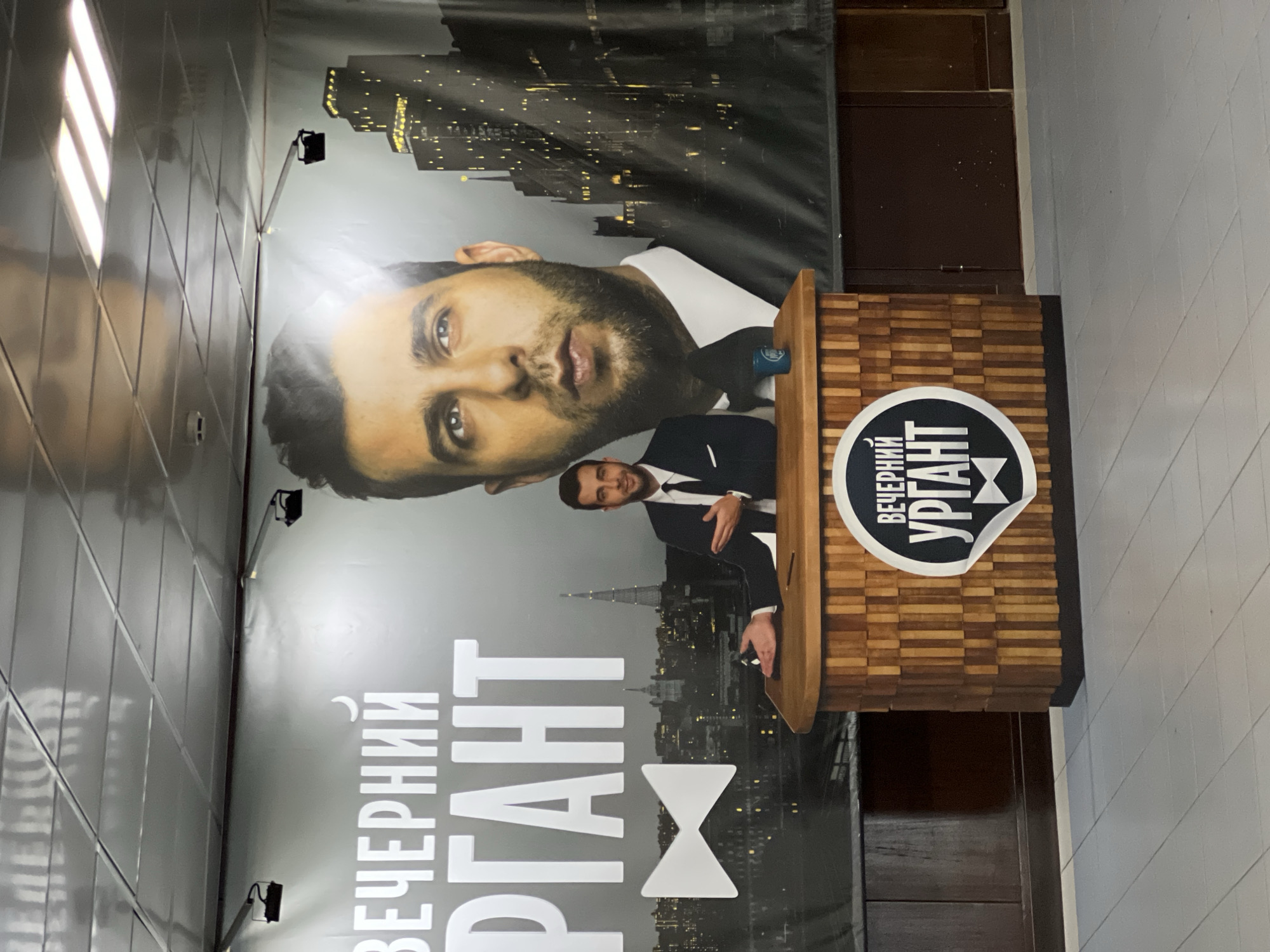 Here is a description of the image:
The image shows an advertisement poster for the Russian late-night talk show "Evening Urgant." The poster features the host, Ivan Urgant, in both a close-up portrait and a full-body shot in a suit. The backdrop depicts a cityscape. A wooden podium with the show's logo is placed in front of the poster. The advertisement is located in an indoor area. Based on the logo and lettering, the poster is clearly in Russian. While Khimki, Russia, is not directly observable in the image, the show itself originates from Russia, so it is possible that the advertisement is located there.
