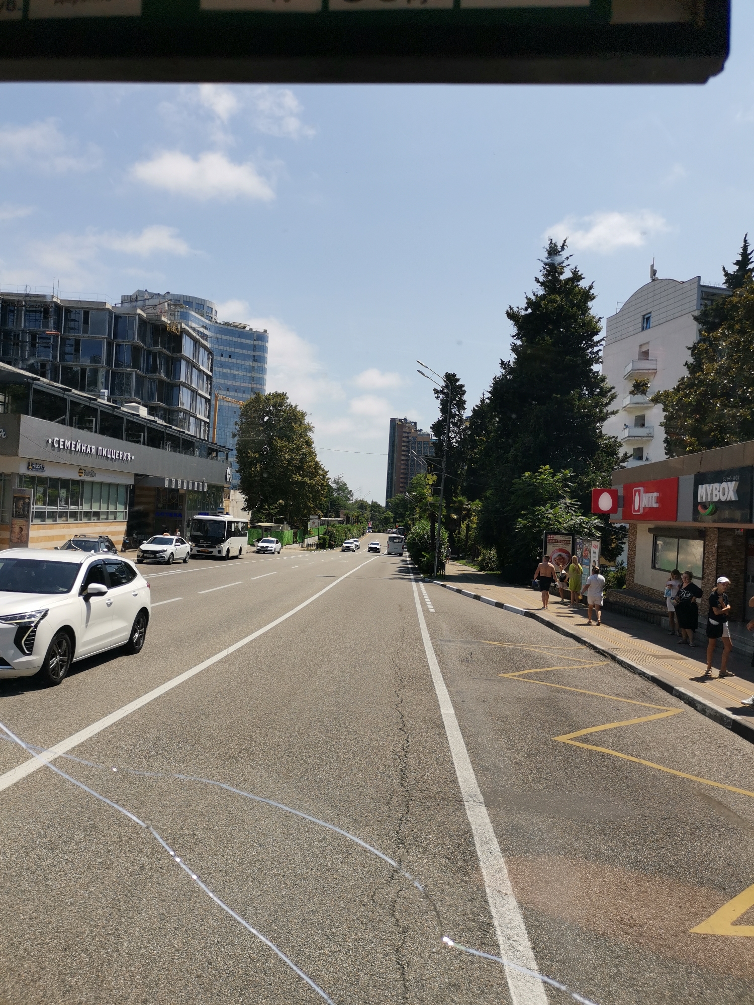 The image depicts a street scene in Sochi, Russia, likely near the coast given the presence of palm trees and the overall atmosphere of a resort town. The perspective is from inside a vehicle, looking forward down a multi-lane road.
On the left side of the road, there is a modern building with a glass facade, under construction in some parts, and partially completed. Below this, a lower structure houses shops, with a prominent sign in Russian that reads "СЕМЕЙНАЯ ПИЦЦЕРИЯ" (Family Pizzeria). A white SUV is parked on the left side of the road in the foreground. Further down the road, several other vehicles, including a white bus and a white taxi, are visible.
The right side of the road features a sidewalk with several people standing and walking. There's a kiosk or small building with signs that appear to be for mobile service providers, including "МТС" (MTC) and "MYBOX". Behind the kiosk and sidewalk, lush green trees and some taller buildings are visible.
The road itself is paved and marked with white lines. Yellow zig-zag markings on the right side of the road suggest a bus stop or waiting area. The sky is blue with scattered white clouds, indicating a sunny day. The overall scene captures a bustling urban environment with elements of commerce, transportation, and ongoing development.