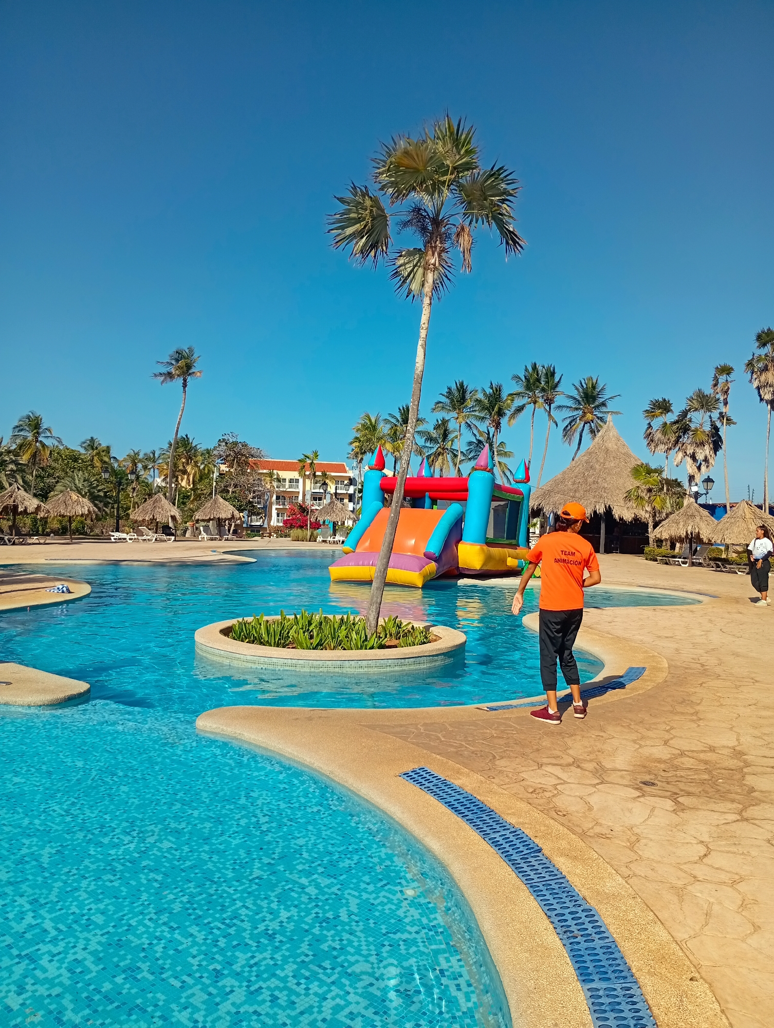 The image captures a vibrant outdoor scene at a resort in Playa El Agua, Venezuela. The focal point is a large swimming pool filled with turquoise water. A small island with greenery is situated in the middle of the pool, and a colorful inflatable water slide is positioned nearby.
A man in an orange t-shirt and dark pants stands near the pool's edge. The t-shirt has the words "TEAM ANIMACION" printed on it. He is wearing a matching orange cap. In the background, another woman is visible, wearing a white shirt and dark pants.
Several thatched-roof structures provide shade around the pool area. Palm trees dot the landscape, and a clear blue sky completes the scene.