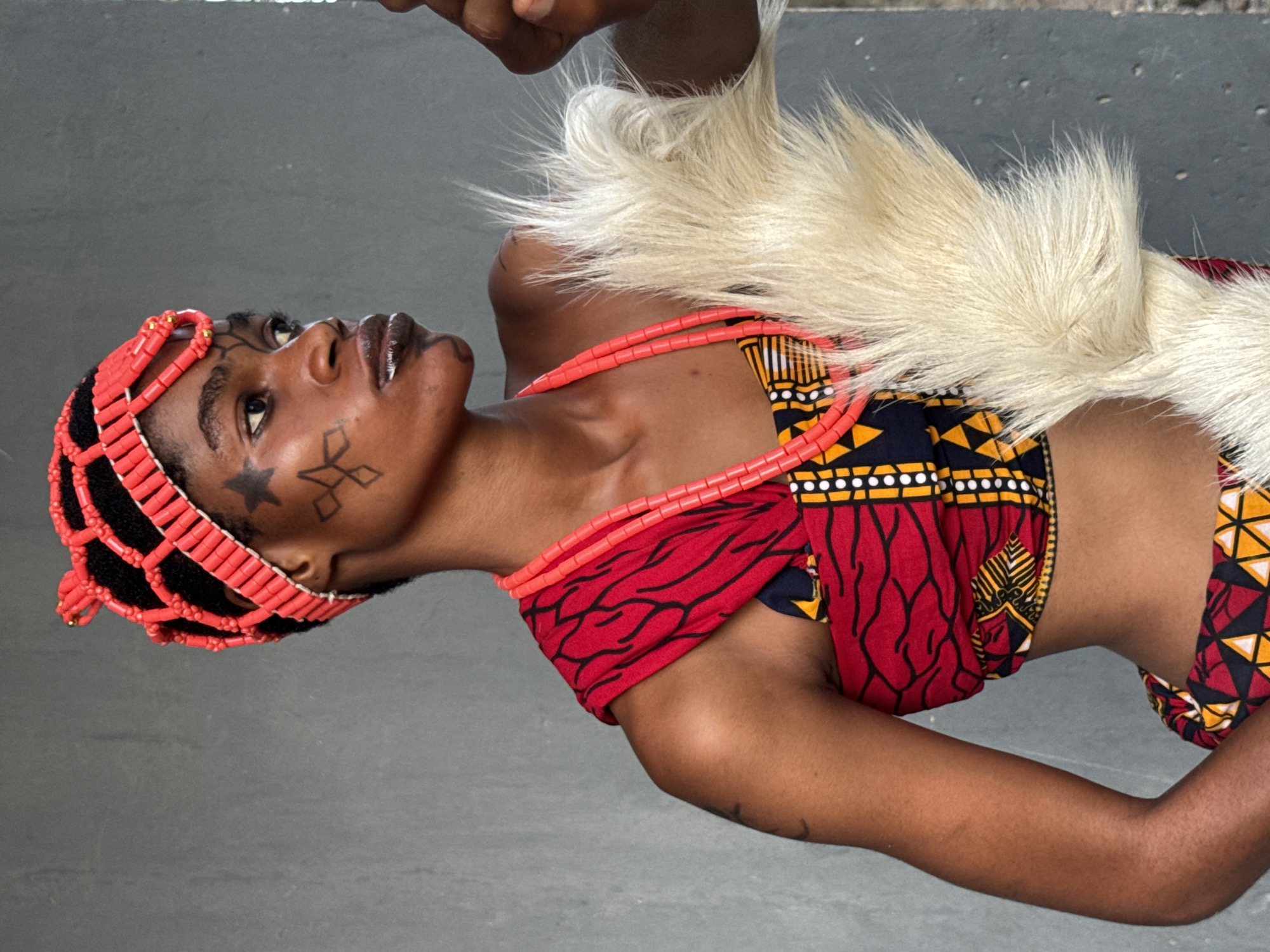 A woman wearing traditional Nigerian clothing is captured in a medium shot against a grey background. She has a headdress made of red beads on her head and a necklace of similar beads around her neck. Her top is a colorful combination of red, yellow, black and white patterns, with a furry white piece draped over her shoulder. Facial markings, including a star and geometric shapes, are visible on her face. The image is taken in Badagry, Nigeria, highlighting cultural attire.