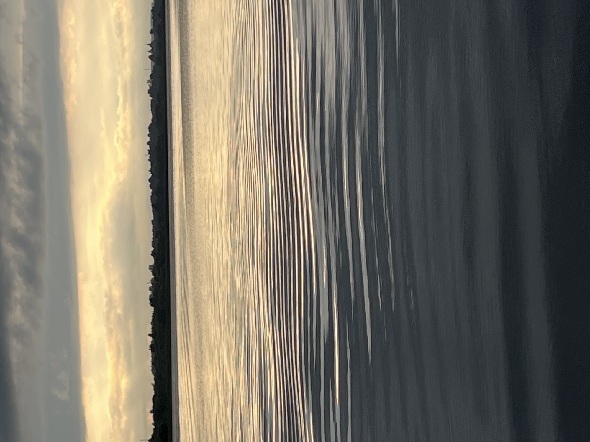 The image depicts a serene body of water with distinct ripple patterns. The water transitions from a lighter, reflective surface in the middle ground to a darker, calmer surface in the foreground. In the background, a silhouetted tree line separates the water from a sky filled with dramatic clouds. A band of warm, golden light streaks horizontally across the middle of the sky, suggesting either sunrise or sunset. The overall impression is one of natural tranquility. The prompt indicates this scene is located in the city of Radchenko, Russia, though this information is not visually verifiable from the image itself.