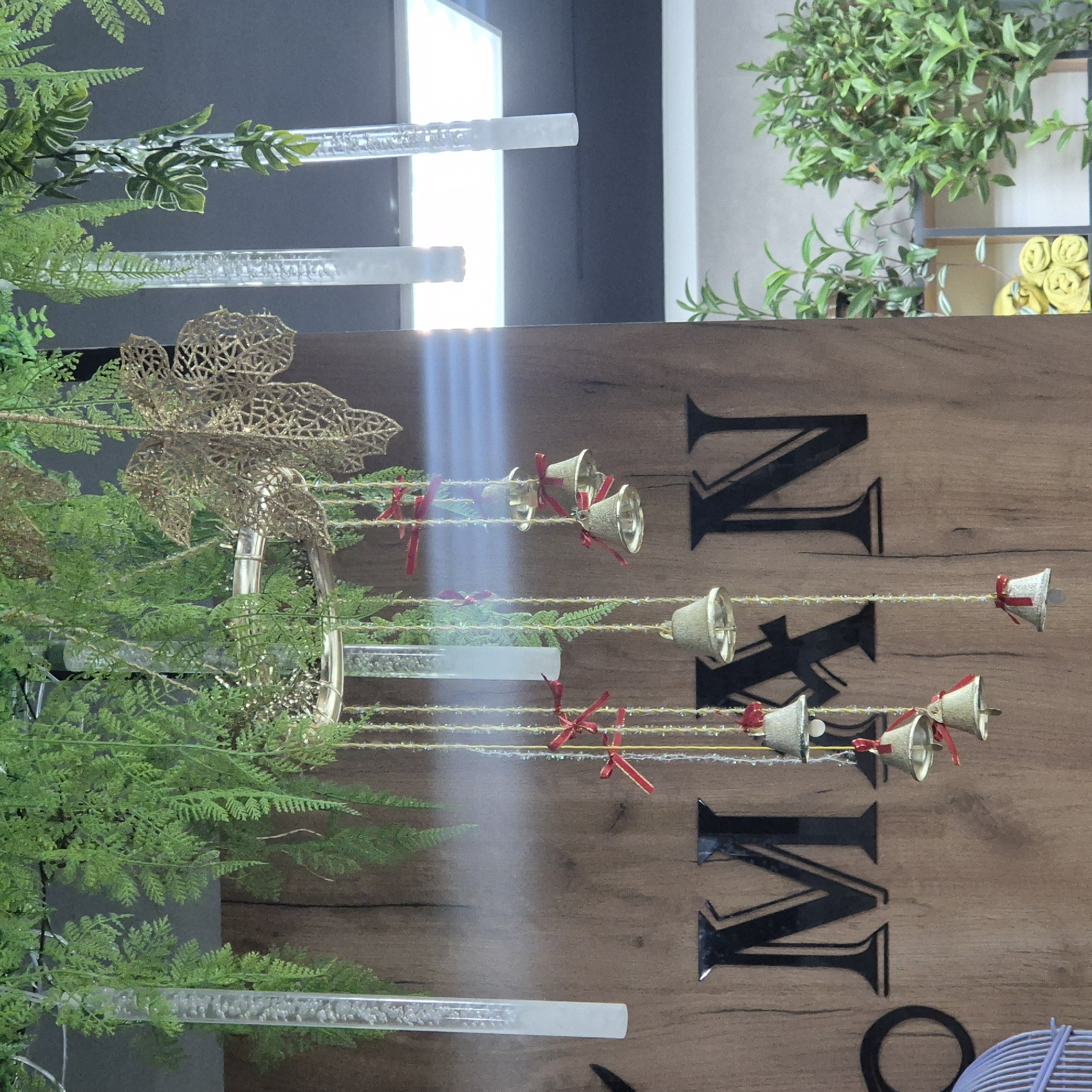 The image shows a decorative arrangement of hanging bells with red bows and fake greenery. The bells are suspended by thin strings adorned with small, clear beads. Behind the decorations, a wooden surface displays large, black, stylized letters that appear to spell "MAN". To the left of the letters, a partial view of a light purple birdcage is visible. The overall scene is likely an interior decoration, possibly in a retail space or a home, given the presence of plants and artificial foliage. There are no people or animals present in the image. The lighting suggests an indoor environment with some bright light sources reflecting off the decorations. The background includes a dark ceiling and a white illuminated panel, possibly a light fixture. There is no indication of the location being in Kokshetau, Kazakhstan.