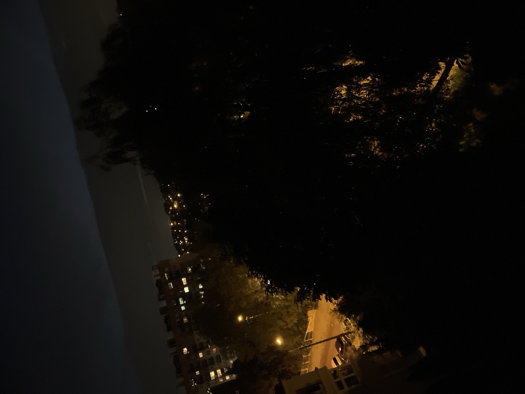 This is a nighttime, high-angle, long shot photograph taken in an urban environment. The image is dominated by darkness, with the exception of artificial lighting from buildings and streetlights. In the background, apartment buildings with illuminated windows are visible, suggesting a residential area. A street with a few parked cars is illuminated by a streetlight. A large, dark tree with visible leaves occupies a significant portion of the foreground, obscuring much of the view. The sky above is dark with a faint, horizontal band of lighter cloud or light pollution. The overall impression is of a quiet, dimly lit city scene at night.