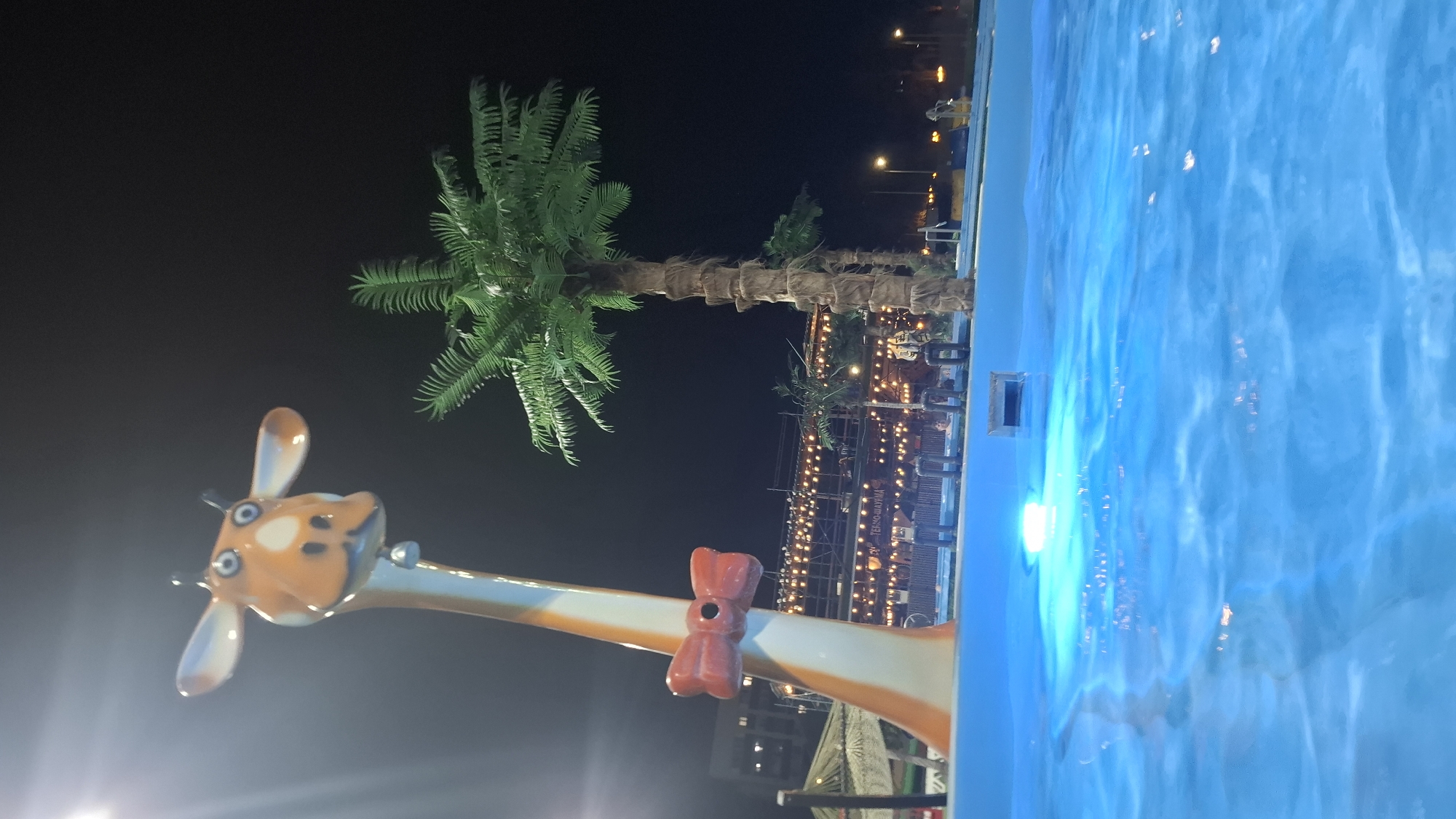 The image shows an outdoor scene at night. The right side of the frame is dominated by a pool of bright blue water, with visible ripples and reflections. Adjacent to the pool is a decorative giraffe statue, featuring a long neck, orange and white coloring, and a red bow. Behind the giraffe, a man-made palm tree stands, adding a tropical ambiance. In the background, there's a structure adorned with strings of lights, possibly a bar or restaurant, with the word "TERMO-MAFINIA" visible. Streetlights and buildings punctuate the dark sky, suggesting an urban setting. The location appears to be in the city of Nizhnekamsk, Russia.