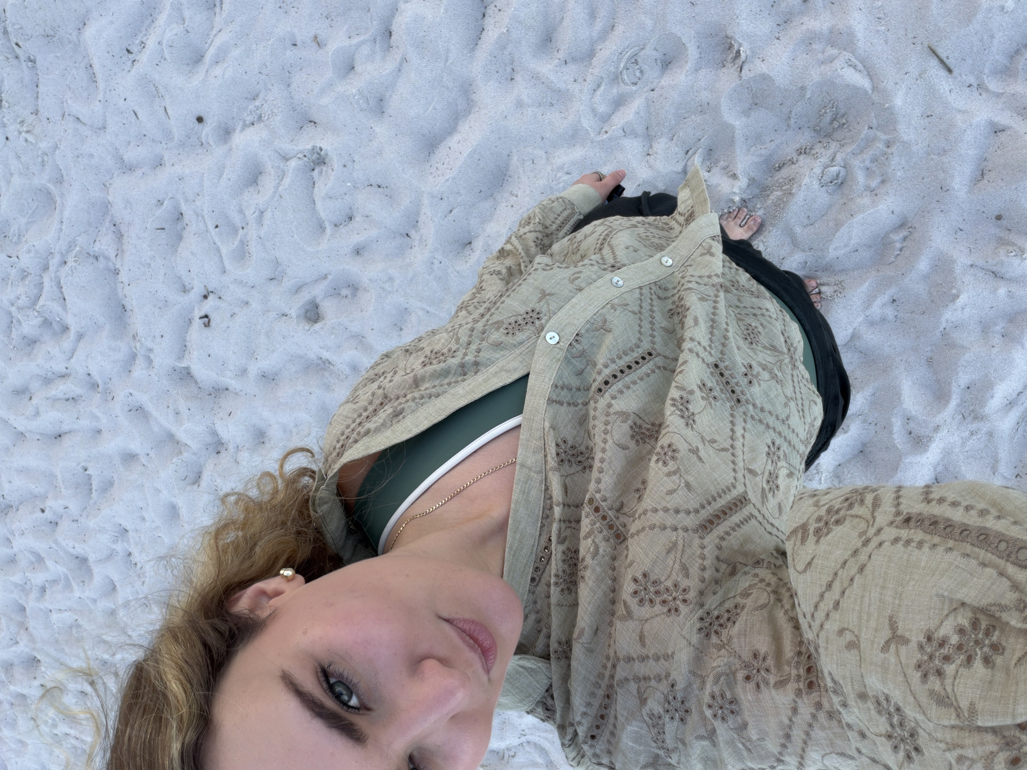 A high-angle selfie shows a woman on a white sandy beach. She is wearing a dark green sports bra, a gold chain necklace, a beige button-down shirt with an embroidered pattern, and dark pants. The location appears to be a beach in Key Biscayne, based on the white sand. The woman is smiling slightly at the camera. Her feet are visible in the background. The sand shows footprints and uneven texture.