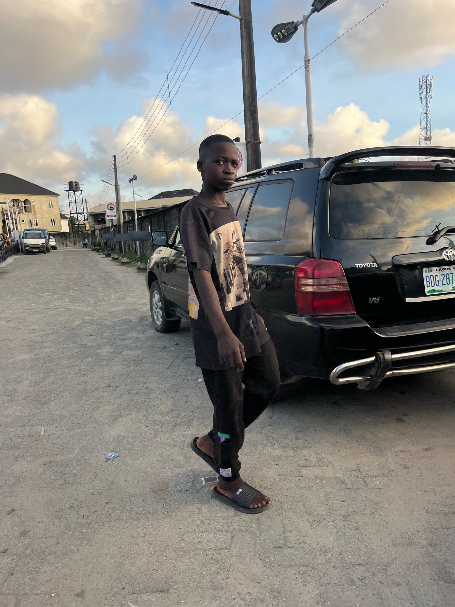 A young Black male stands on a cobblestone street in Ado, Nigeria, next to a black Toyota SUV. He is wearing a black t-shirt with a graphic design on the front, black pants, and black sandals. His left hand is resting on his right leg. The SUV's license plate reads "BDG-287" and has a Lagos sticker. In the background, there are buildings, a water tower, and utility poles with wires. The sky is cloudy with patches of blue. A cell tower is visible in the distance. There is a discarded item on the ground near the young male's feet.
