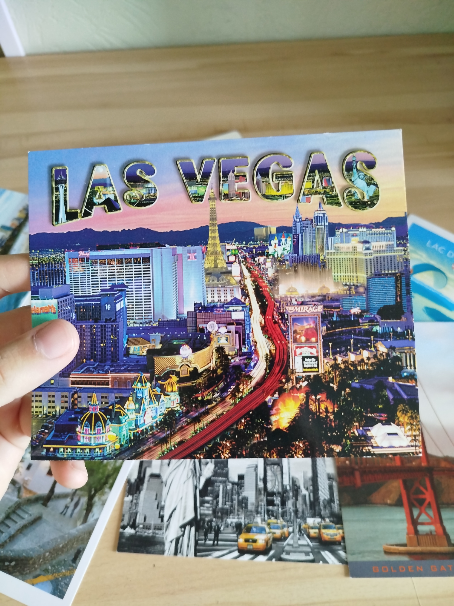 The image shows a postcard with the embossed text "LAS VEGAS" across the top. The postcard depicts a nighttime aerial view of the Las Vegas Strip, featuring illuminated hotels, casinos, and a highway. In the foreground, a person's thumb and index finger are visible, holding the postcard. Partially visible to the left and below the main postcard are sections of other postcards or photos. One appears to be a black and white cityscape with yellow taxis. Another shows a bridge, with the text "GOLDEN GATE" at the bottom. The overall scene is one of a collection of travel-related memorabilia displayed on a wooden surface. The location is not Varna, Bulgaria; it is Las Vegas, Nevada, USA.