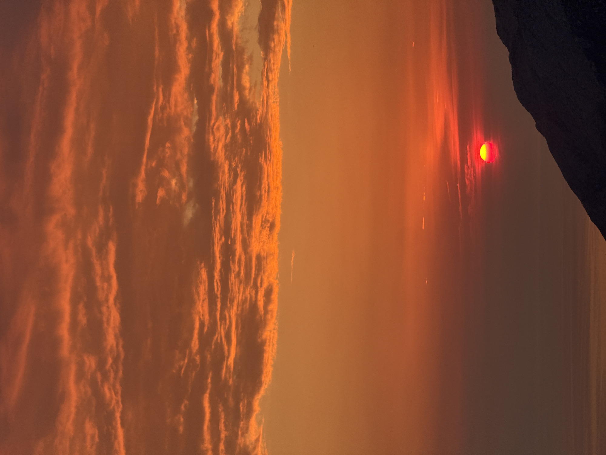 The image captures a dramatic sunset over the Adriatic Sea, with the sun a vibrant red disc partially obscured by haze, low on the horizon. The sky is ablaze with hues of orange and red, with streaky clouds overhead adding texture to the scene. The silhouette of a dark, mountainous landmass dominates the lower right portion of the frame, providing a stark contrast to the fiery sky. The sea is visible in the bottom left, reflecting the dim light of the setting sun. The location is identified as Gluhi Do, Montenegro, a coastal settlement known for its scenic beauty. There are no people or specific objects of interest visible in the image, only the natural spectacle of the sunset over the sea and mountains.
