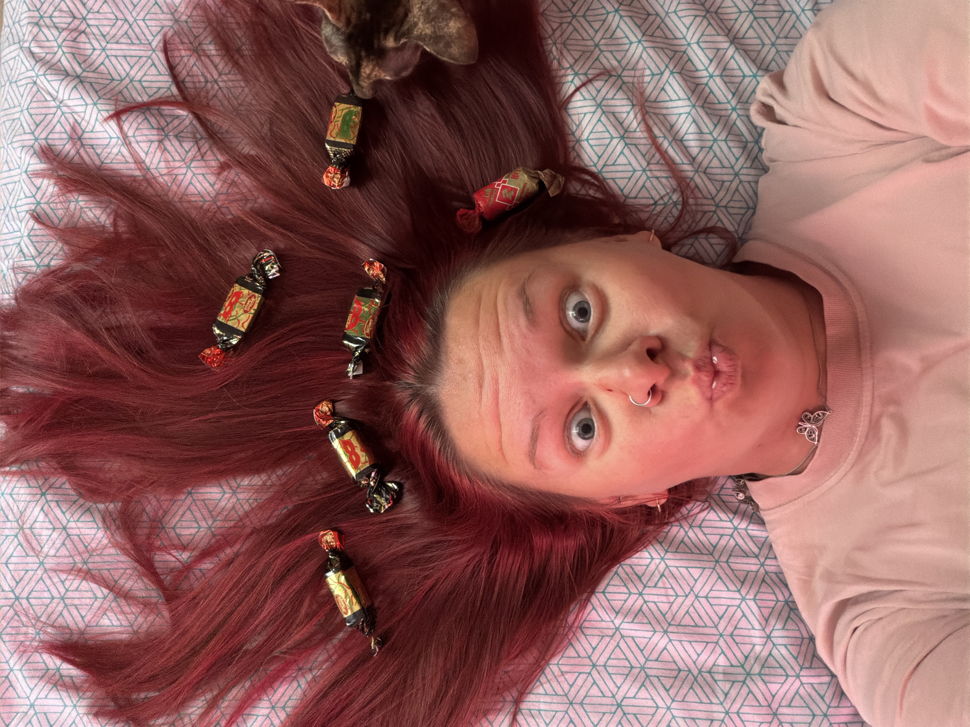 The image is a close-up, high-angle shot of a person with long, red hair lying on a bed with a geometrically patterned pink and teal bedsheet. Six individually wrapped candies are placed sporadically within the person's hair. A cat appears at the top left of the frame, its head partially visible. The person has fair skin and is wearing a light pink sweater. They are looking directly at the camera with wide eyes and pursed lips. They have a nose ring and a necklace with a butterfly pendant. The setting cannot be identified as Chaikovsky, Russia, based on the visual information provided.