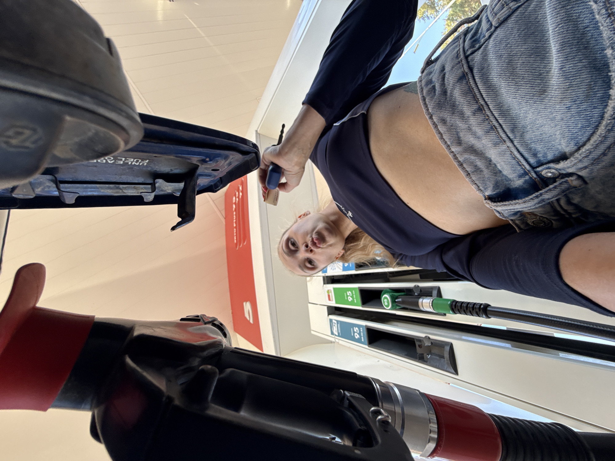 A woman is refueling a blue MG car at a gas station in Chaikovsky, Russia. The pump shows 95 octane gasoline. The woman is holding a blue object in her hand. She is wearing a navy long-sleeve shirt and denim shorts. The scene is lit by natural daylight.