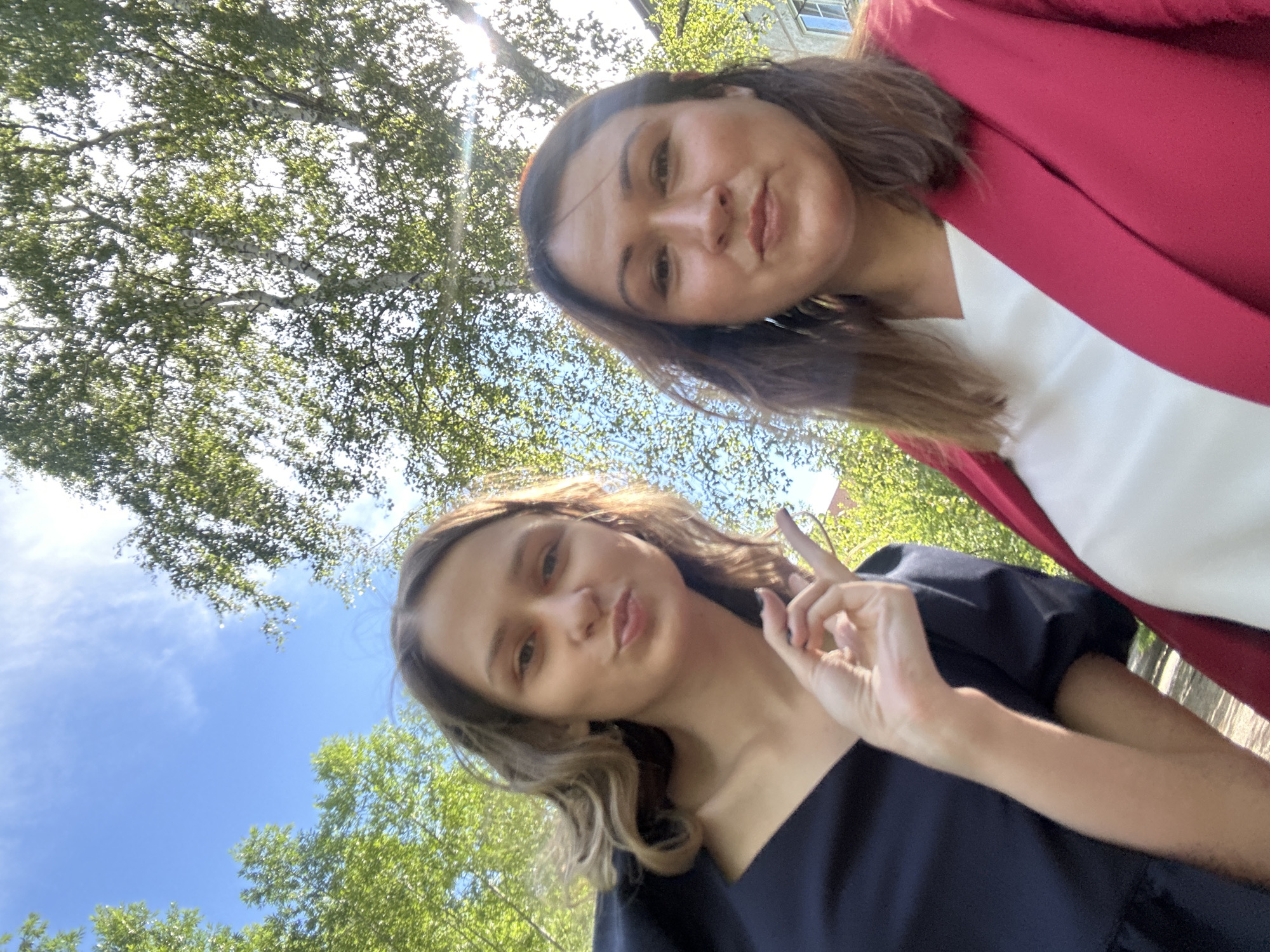 Two women are taking a selfie outdoors. The woman on the left is wearing a dark blue dress and has her hair down, wavy, and blonde. She is holding up her hand with her index and thumb forming a circle, and her lips are pursed. The woman on the right has brown hair and is wearing a white top under a red jacket. Both women are smiling faintly. They are positioned in front of a bright blue sky with sunlight filtering through green tree leaves, suggesting a sunny day. The background also shows some buildings. The scene appears to be in a park or a residential area with trees. There are no specific indicators of the city being Omsk, Russia.