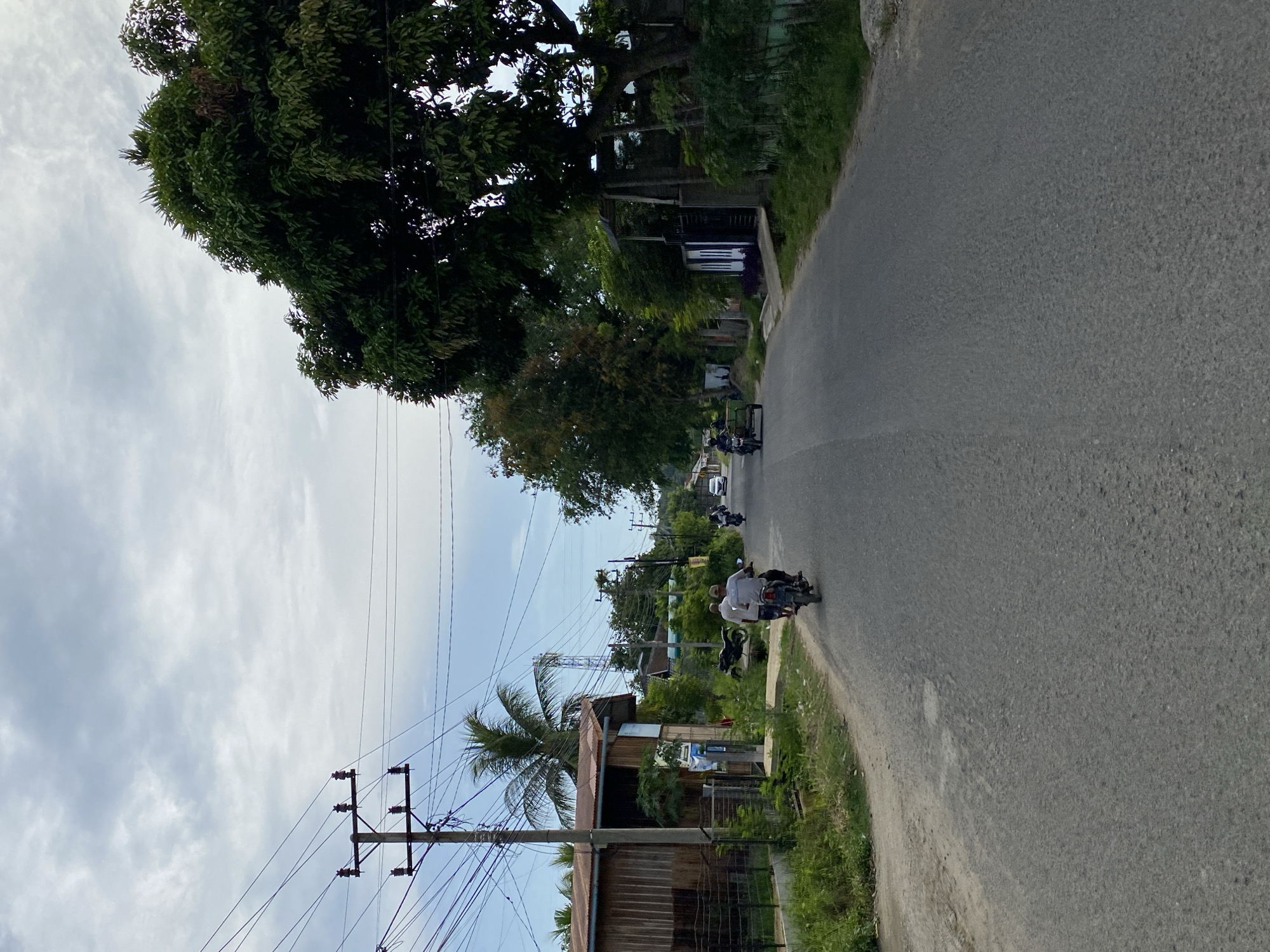 This image depicts a road in Lhokseumawe, Indonesia, likely a residential or semi-rural area. The road surface is asphalt, showing signs of wear and tear with some patching. The environment is characterized by lush green vegetation lining the road, with several trees and palm trees visible. A tall wooden utility pole with multiple power lines stretches across the left side of the frame, suggesting the presence of electricity infrastructure.
On the road, several vehicles are present. Two motorcycles are prominently featured. The first, in the foreground, has two occupants, a driver and a passenger, both wearing light-colored shirts. The second motorcycle, further down the road on the right side, also appears to have two occupants, one of whom is operating the vehicle. Another motorcycle with a sidecar is visible on the right side of the road, also with at least one occupant. In the distance, on the right side, a white car is visible.
Buildings are present on both sides of the road. On the left, a simple wooden structure with a corrugated metal roof is visible, partially obscured by vegetation. On the right, houses with fences and dense foliage are present. The sky is cloudy, with a mix of gray and lighter patches, indicating an overcast or partly cloudy day. The overall impression is a typical scene of daily life and transportation in a community in Lhokseumawe.