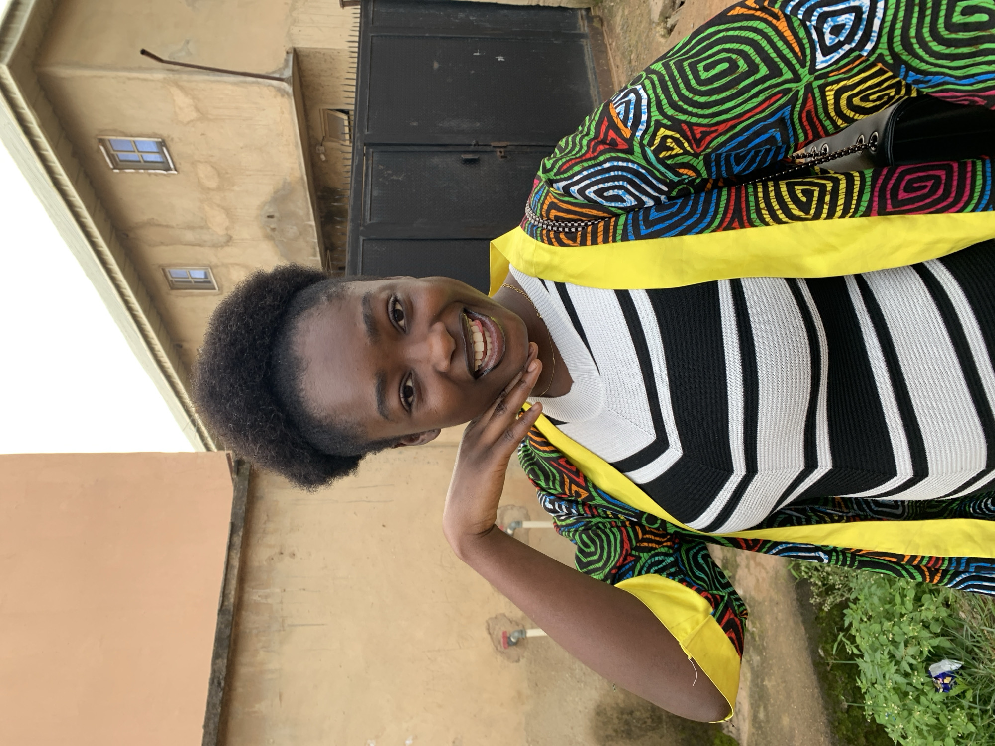 A young woman of color with dark, curly hair is posing for a photo in front of a beige building with a black metal gate. She is wearing a black and white striped shirt and a colorful, patterned jacket with yellow trim. Her left hand is resting on her cheek, and she is smiling with her mouth open, showing her teeth and tongue. The background shows the exterior of a building with windows and a roofline, as well as some green foliage and a discarded piece of paper on the ground. The scene appears to be outdoors, likely on a street or in a courtyard in Rafin Par, Nigeria.