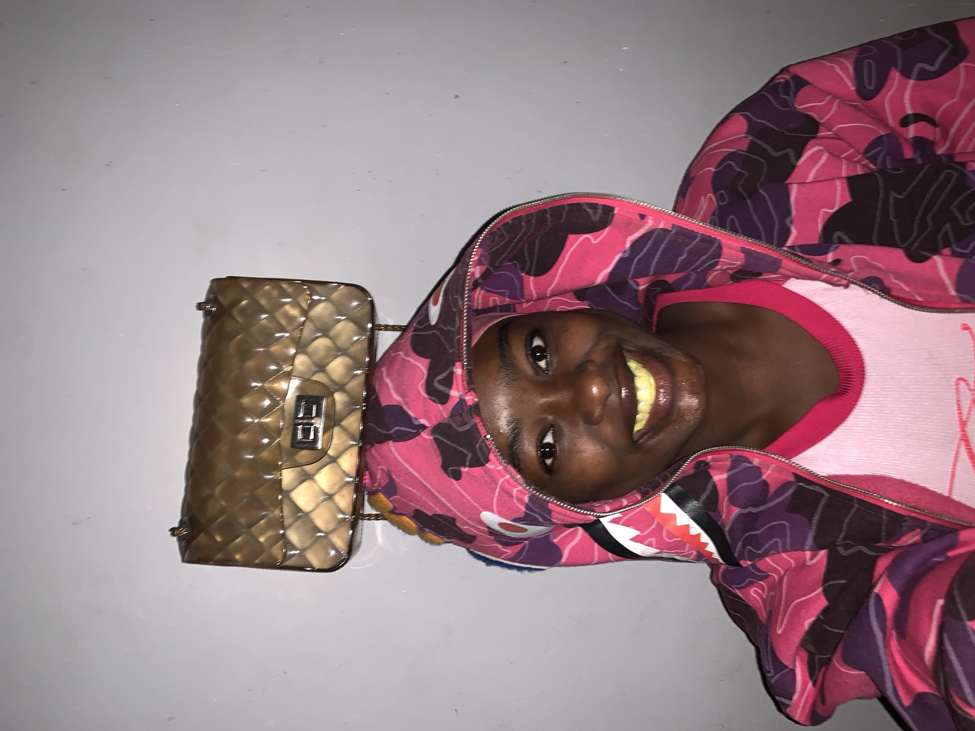 A young Black person wearing a pink and purple camouflage hoodie and a pink t-shirt is posing for a picture. A gold quilted purse is lying on a grey surface beside them. The location is not specified as Rafin Par, Nigeria.