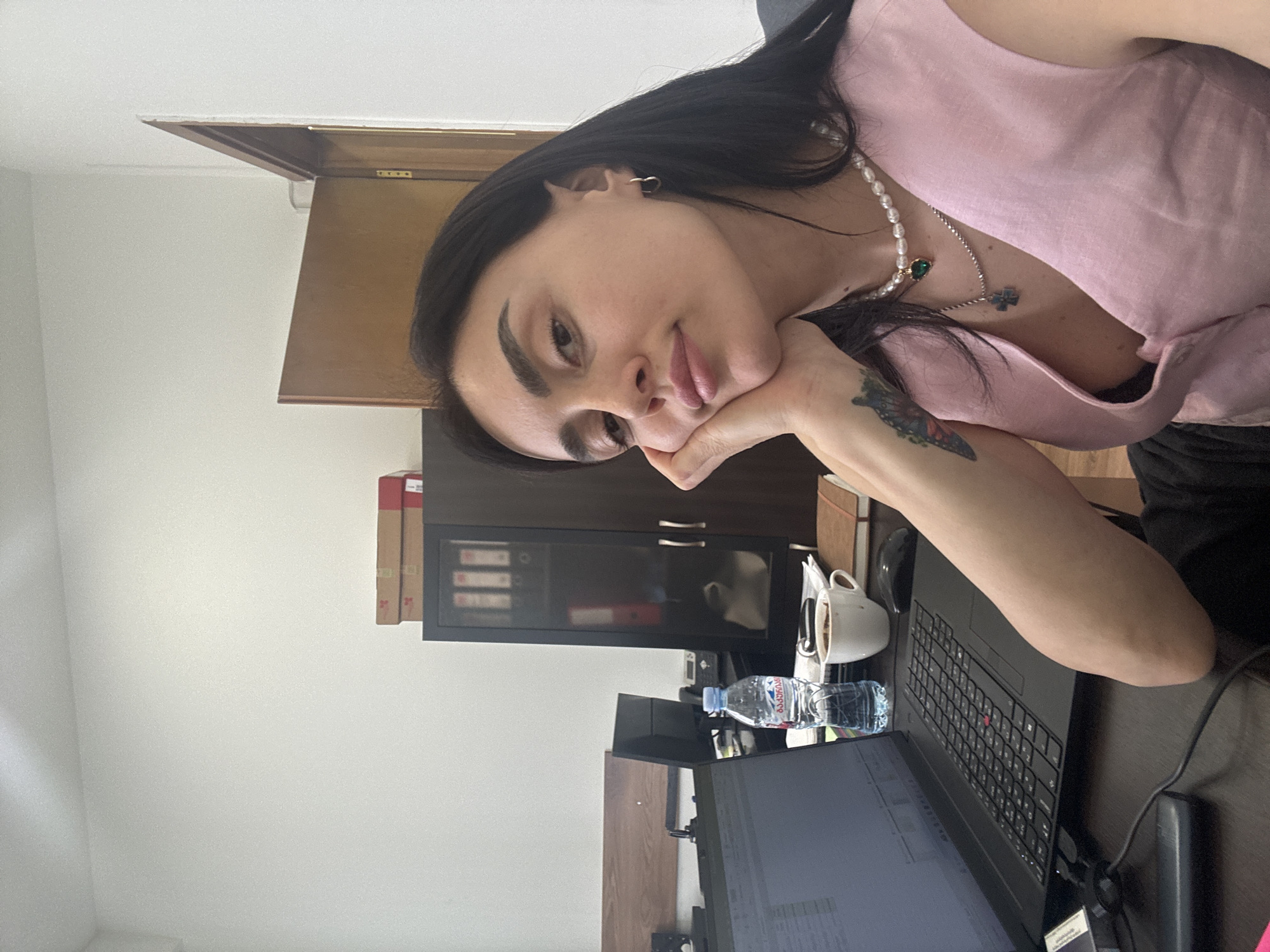 The image shows a woman sitting at a desk. She has dark hair and is wearing a pink top and a pearl necklace. She is resting her chin on her hand, and her gaze is directed towards the camera.
On the desk, there is a laptop, a water bottle with Georgian text on the label, a coffee cup, and other desk items. Behind the desk, there is a cabinet and a door. The wall is white.
The water bottle's label indicates that the image was taken in Georgia. While it's impossible to confirm the exact city from the visual evidence alone, based on the context of the prompt, it is most likely Tbilisi, Georgia.