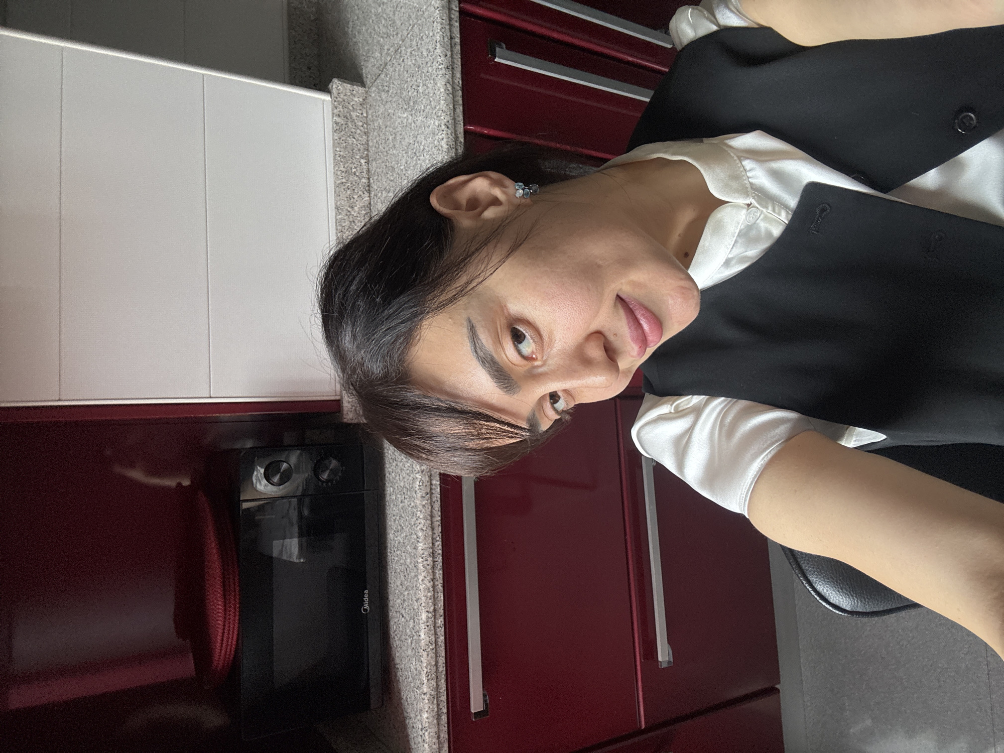 A person with dark hair and fair skin is indoors, possibly in a kitchen. The person is wearing a white collared shirt and a black vest. They have an earring in their left ear. Behind the person are red cabinets with silver handles, a white wall, and a microwave. The location is not identifiable as Tbilisi, Georgia from the image.