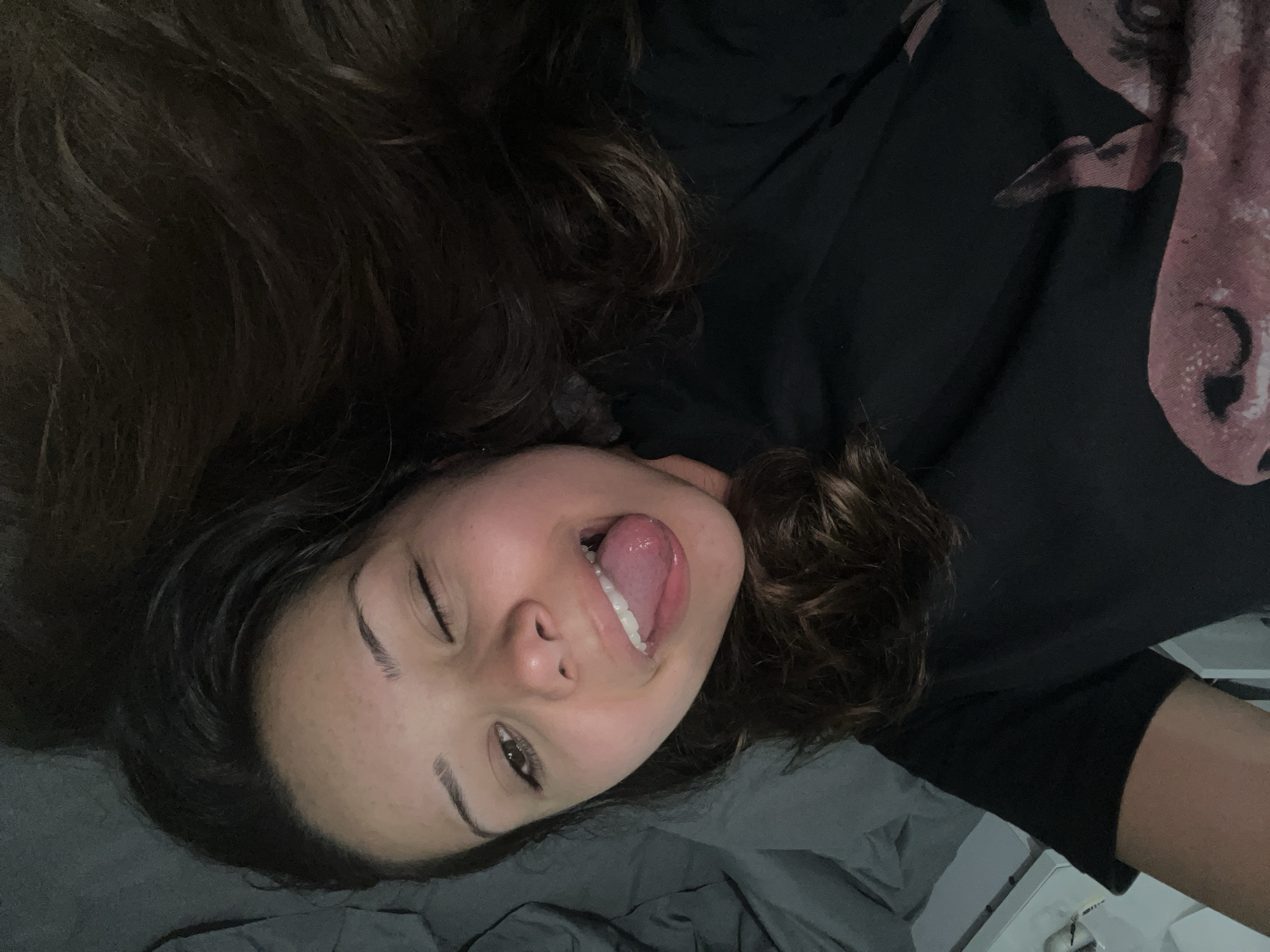 A young woman with dark, wavy hair is lying on her side on a bed with gray bedding, looking up at the camera. She is wearing a black t-shirt with a pink graphic design on it. Her eyes are partially closed in a wink, and her tongue is sticking out, forming a playful expression. The lighting suggests an indoor setting, possibly a bedroom. There is no identifiable information about the specific location within Zlatograd, Bulgaria.