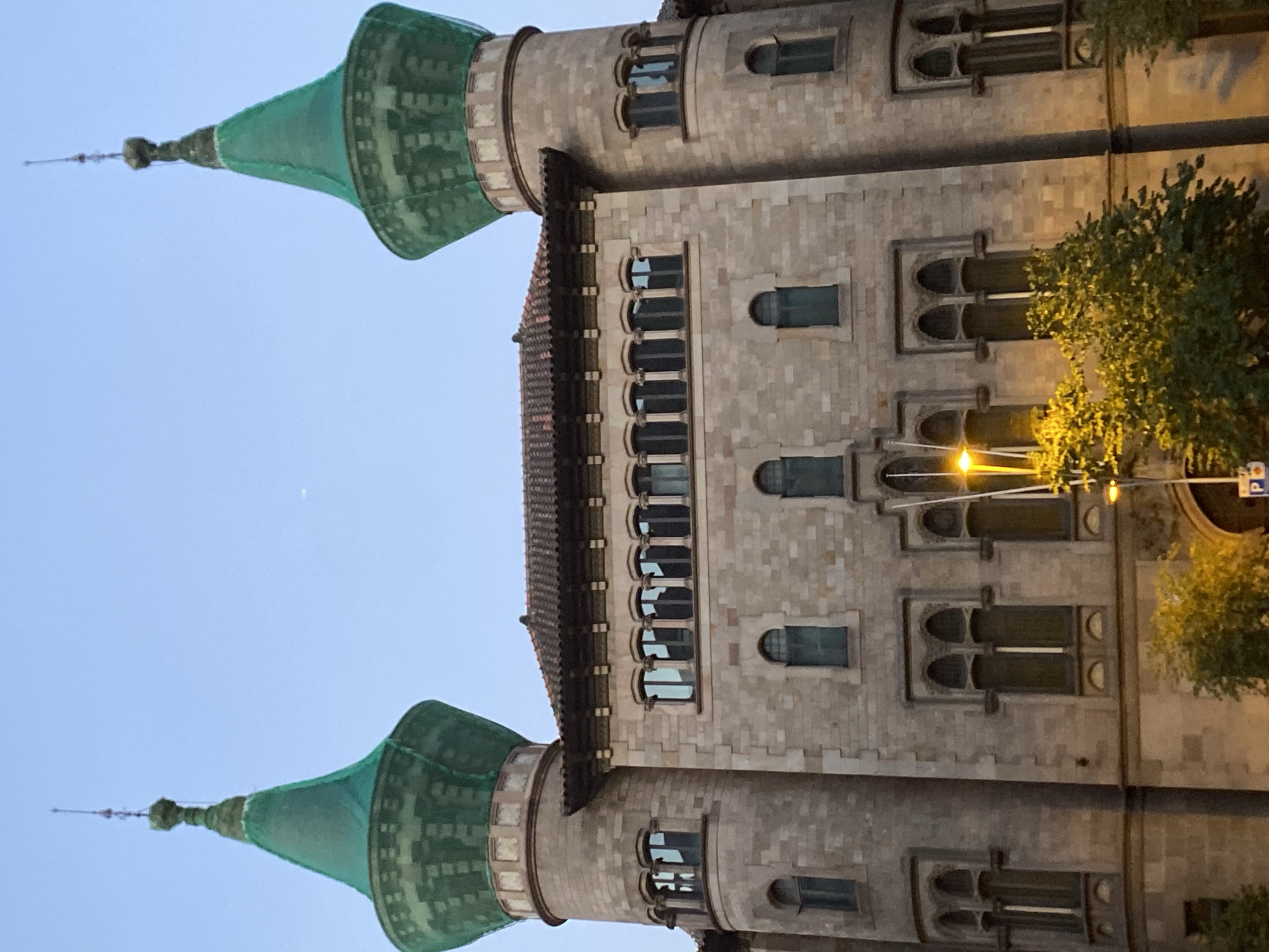 The image shows a building with two prominent conical green towers, each topped with a spire. The building itself is constructed of stone blocks and features a tiled roof. It has numerous windows, some of which are arched and others are rectangular. The overall architectural style appears to be historic. The lighting suggests it is either dusk or dawn, with a clear sky above. In the foreground, there are some trees and a street lamp that is illuminated, casting a warm glow on the lower part of the building and the foliage. The building is located in the L'Eixample district of Barcelona, Spain.