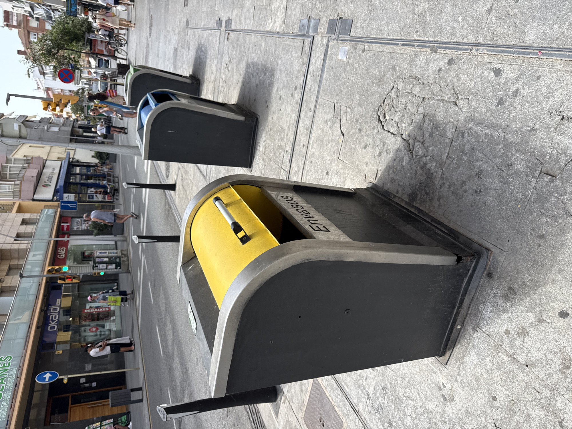 The image shows a street scene in Blanes, Spain. In the foreground, there are three large, modern trash receptacles, one of which is primarily yellow and labeled "ENVASOS". The receptacles are placed on a paved sidewalk next to a street with traffic. In the background, there are various shops and businesses, including "OKAI", "GAES", and "Condis". Pedestrians are visible walking along the sidewalk and crossing the street. The overall environment appears to be a commercial district in an urban setting on a sunny day.