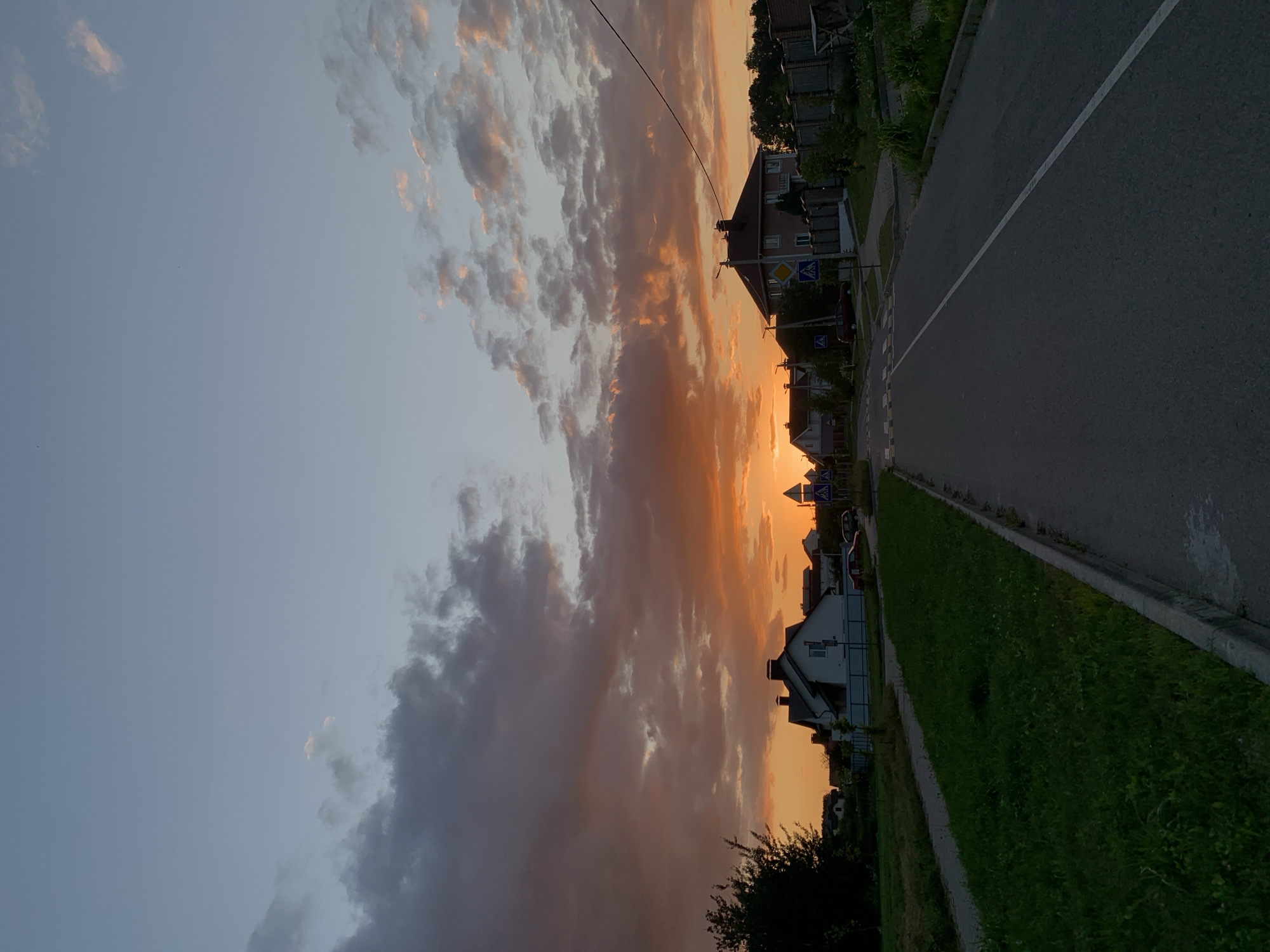 The image depicts a street in Minsk, Belarus, during a sunset. The sky is filled with dramatic clouds painted in hues of orange, pink, and purple, transitioning to a soft blue at the top. Several houses with pitched roofs are visible on either side of the asphalt road. The road has a white dividing line and a pedestrian crossing. Green grass lines the side of the road, and some trees are present. Traffic signs and streetlights are also visible. The overall atmosphere is serene, with the setting sun casting a warm glow over the suburban landscape. There are no people or animals visible in the image.