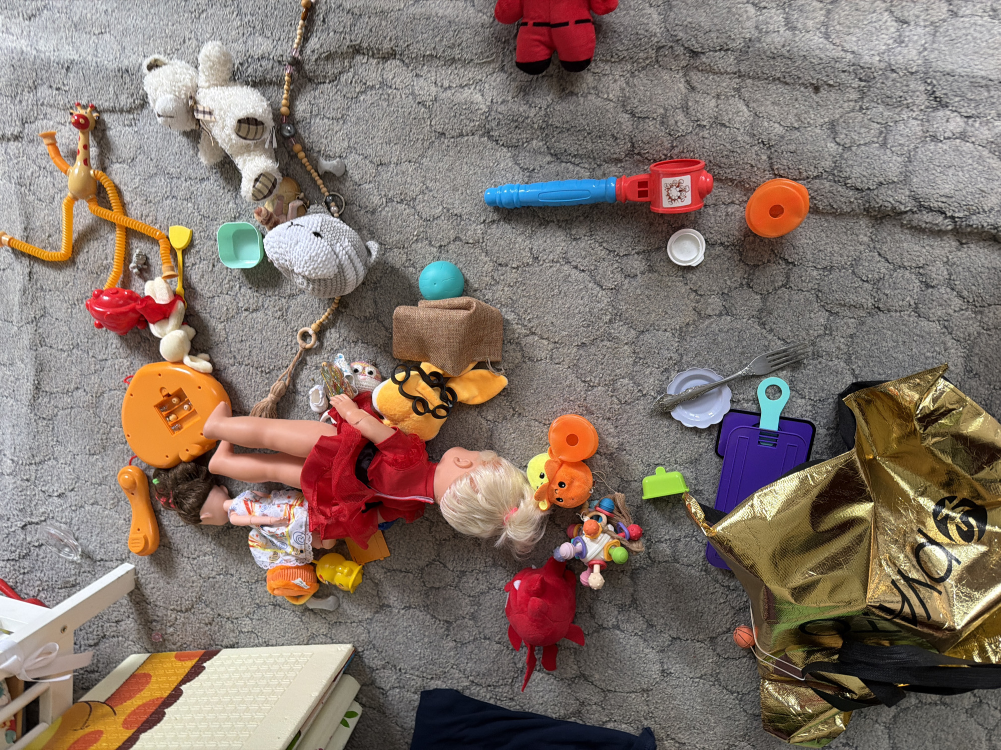 The image displays a collection of children's toys scattered across a gray, carpeted floor. Among the items are a doll with blonde hair wearing a red dress, a stuffed white bear, a plush red monster-like toy, and several plastic toys of various colors and shapes, including what appears to be a toy cell phone, a toy gun or blaster, and a small orange ball. There are also some craft items, such as beads and a golden bag with black lettering. The lighting suggests an indoor setting. There is no information in the image to indicate a specific location in Moscow, Russia.