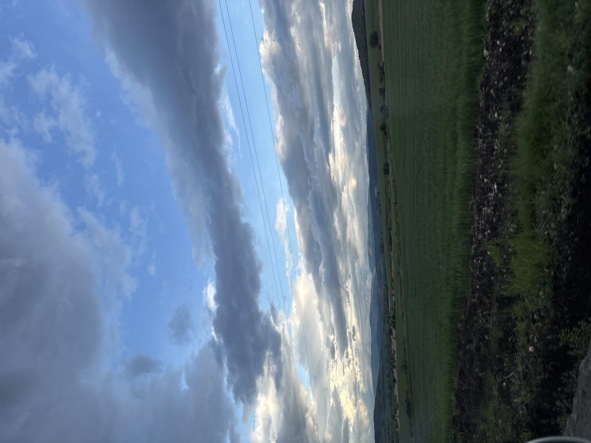 The image shows a landscape near Gorna Malina, Bulgaria. The sky is partly cloudy with blue patches visible between the gray and white clouds. Power lines stretch across the sky. A green field extends into the distance, meeting a tree-lined horizon where a low mountain range is discernible. Along the edge of the field is a darker patch of earth with vegetation growing on it.